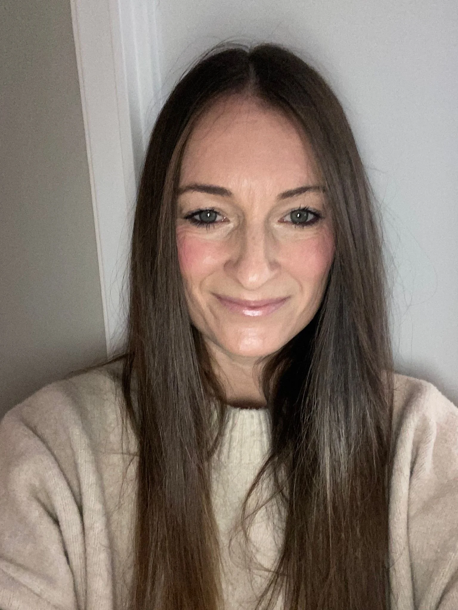 A woman with long brown hair, light makeup, and a beige sweater, smiling in front of a white wall.