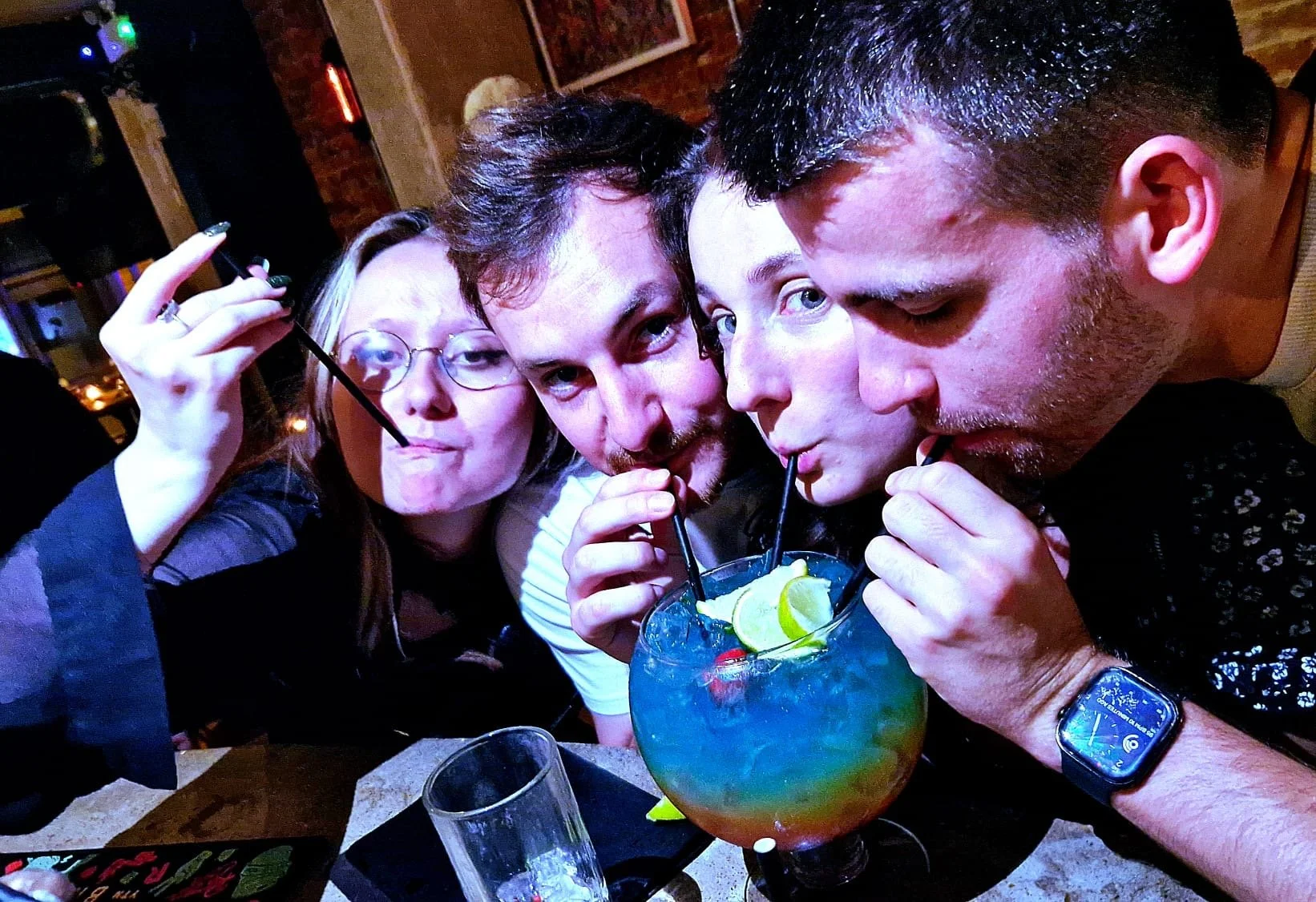 Four friends sharing a large colourful cocktail with lime slices, sipping through straws in a party setting.