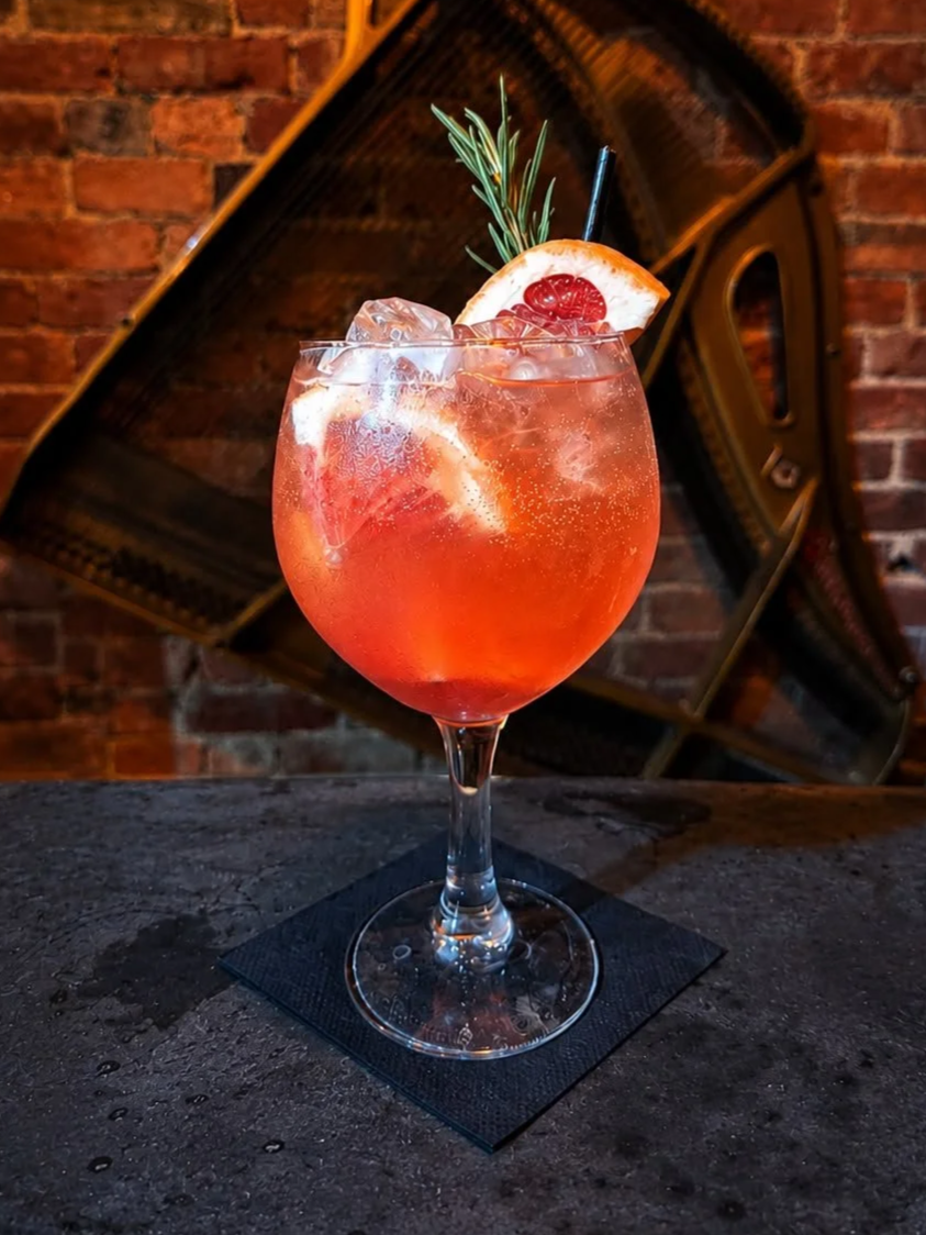 A pink cocktail with ice cubes garnished with a lemon slice, raspberry, rosemary sprig, and straw, served in a large wine glass on a black napkin with a brick wall and piano in the background.