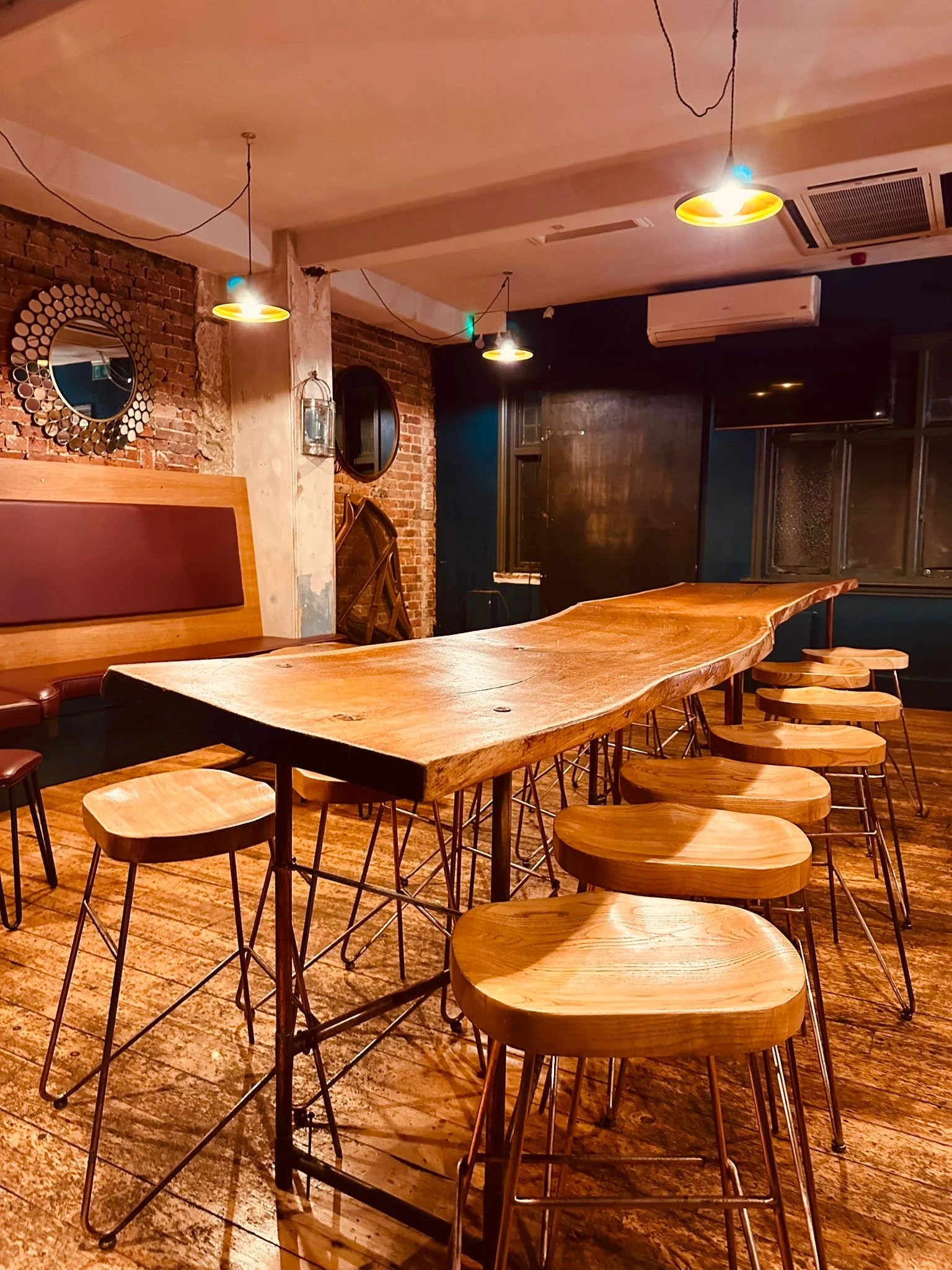 Interior of a bar or restaurant with a long wooden communal table, wooden stools, brick walls, and decorative mirrors on the wall.