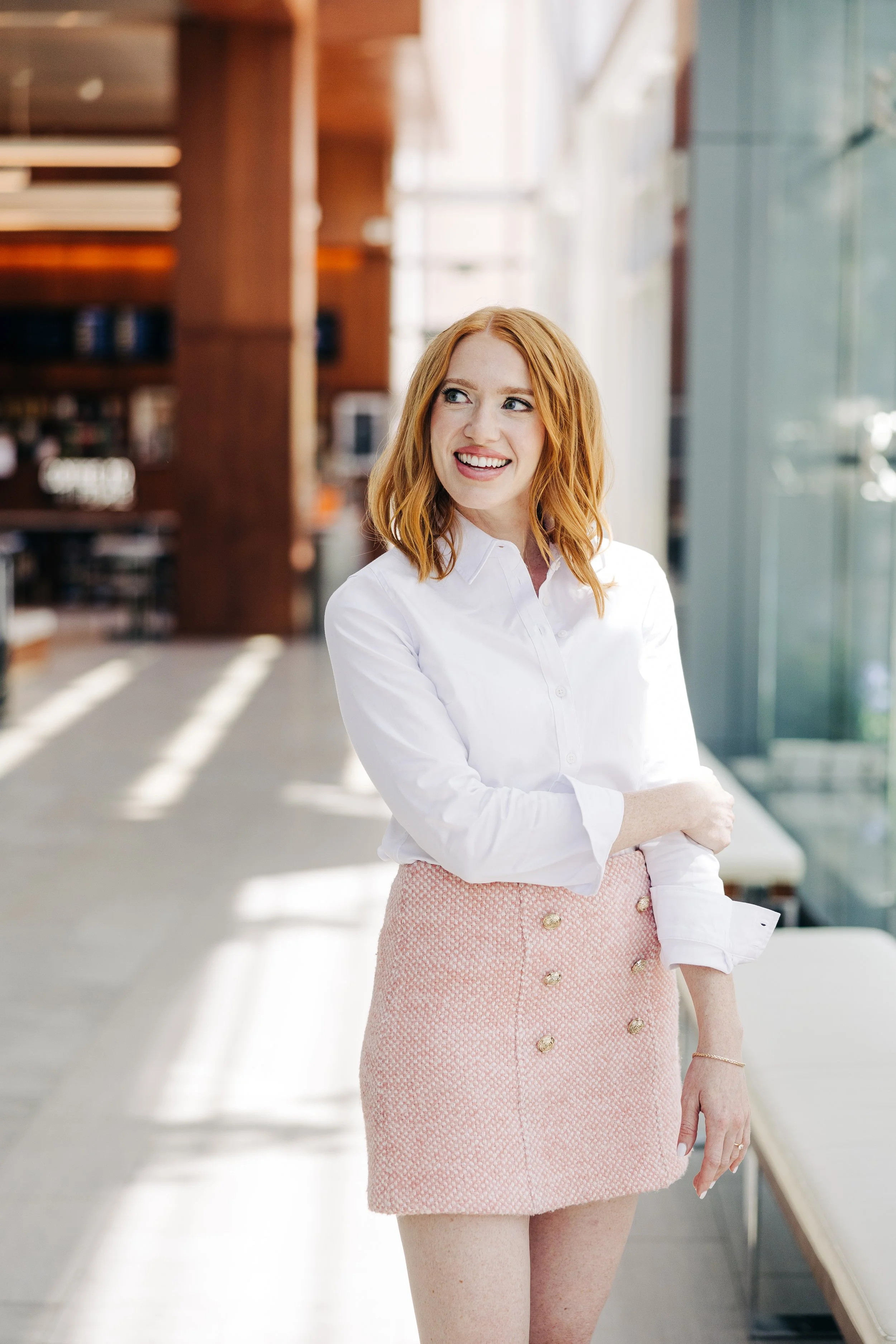 A woman with red hair wearing a white shirt and pink skirt standing inside a modern building with large glass windows.