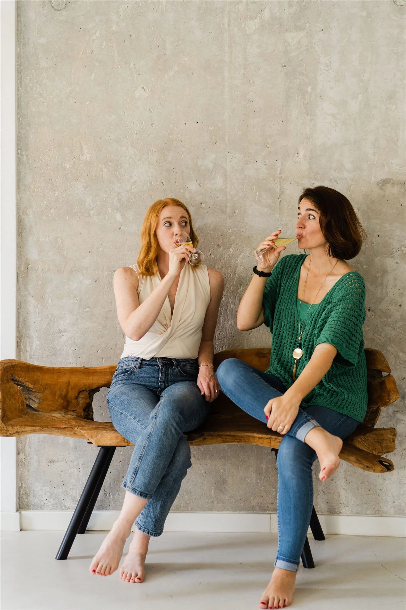 Two women sitting on a wooden bench, drinking from small glasses, against a concrete wall background.