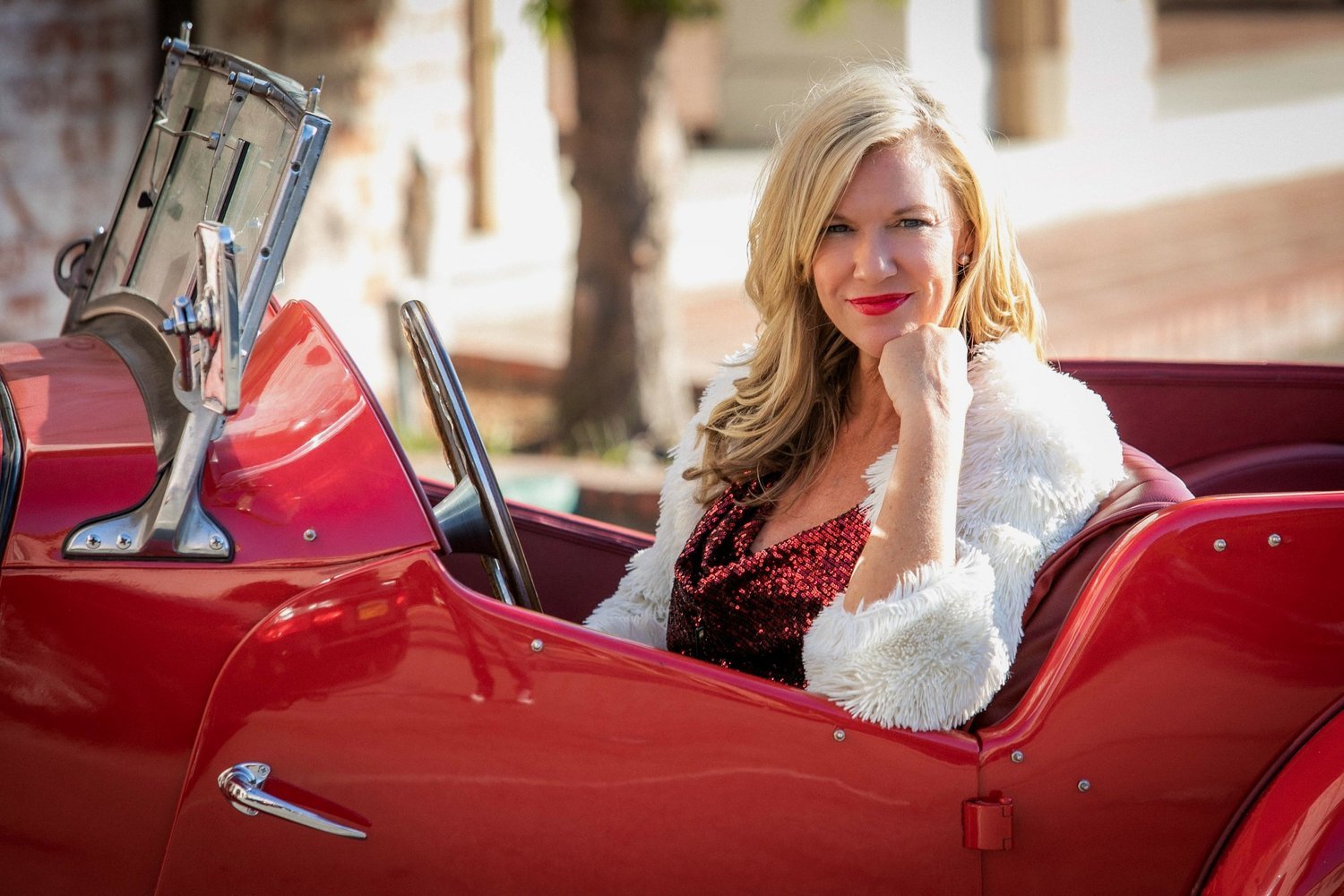 A woman with blonde hair, wearing a white furry jacket and red lipstick, sitting in a red vintage convertible car with a blurred outdoor background.