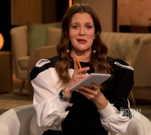 A woman with long brown hair, wearing a black and white dress with puffy sleeves, sitting on a beige couch, holding an orange pen and a notepad, speaking and gesturing with her right hand in a talk show setting.