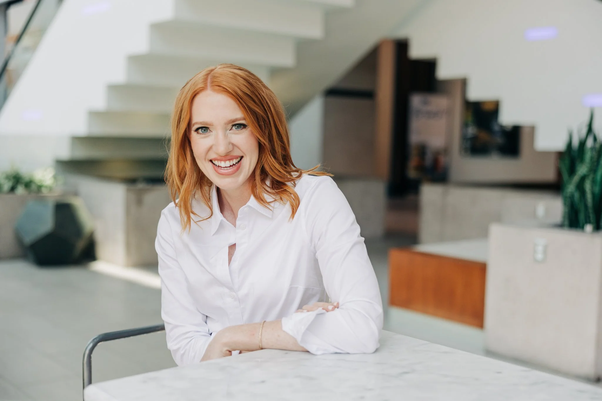 A woman with red hair and a white shirt sitting at a marble table, smiling in a modern interior space.