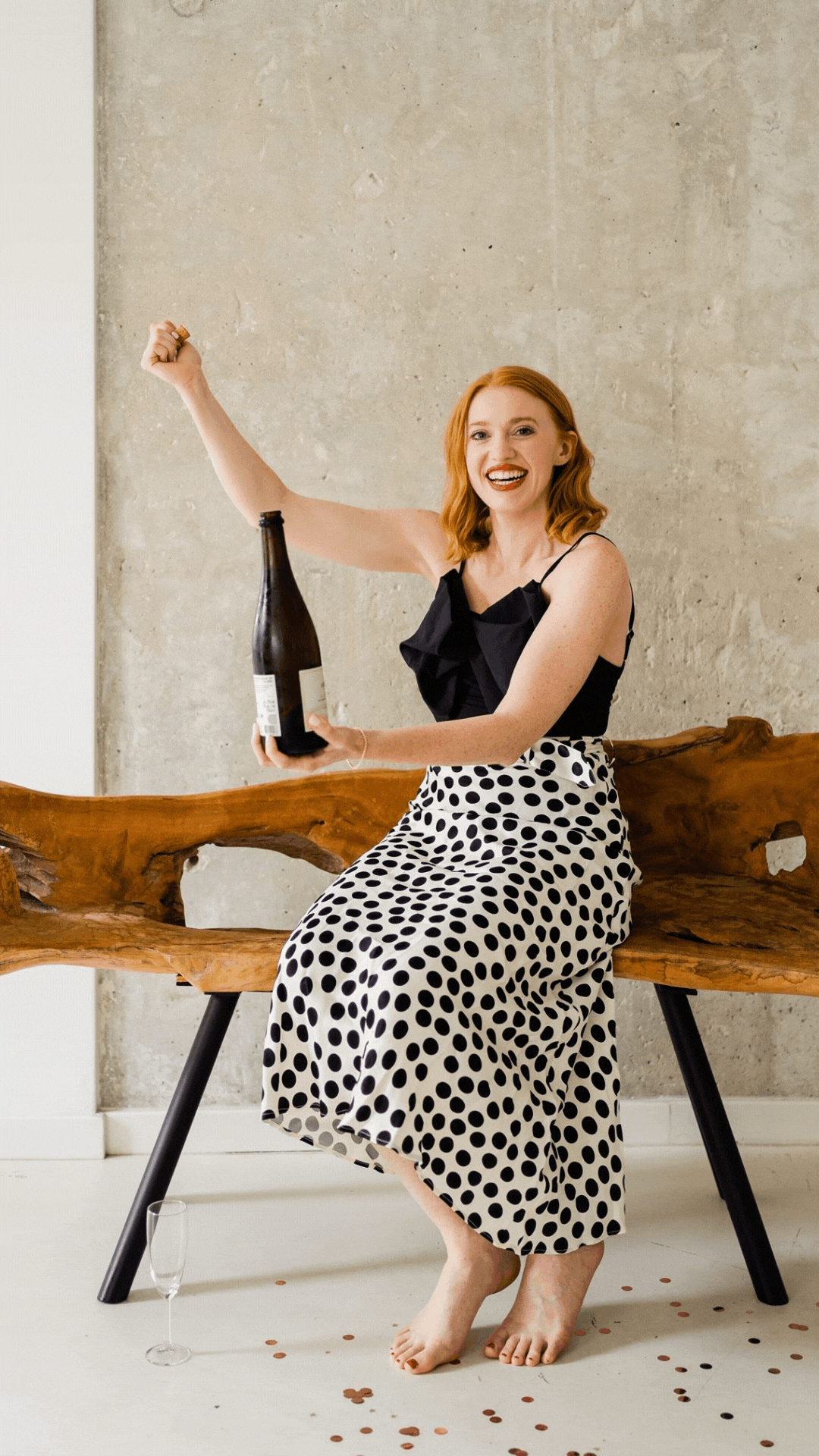 A woman with red hair and a big smile, sitting barefoot on a wooden bench, holding a dark bottle and celebrating with scattered confetti and an empty champagne glass on the floor.