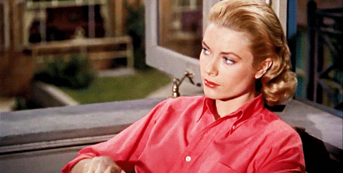 A woman with blonde hair wearing a pink shirt sitting at a table indoors with a window in the background.