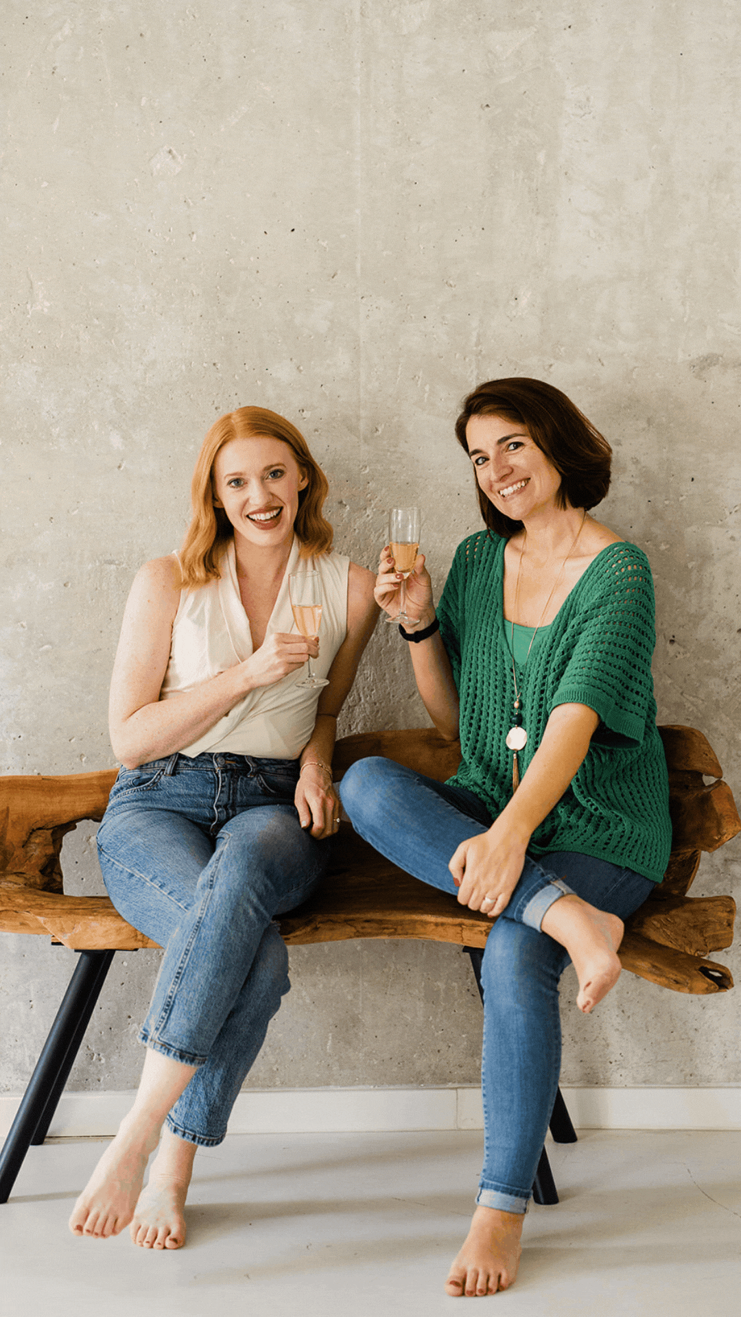 Two women sitting on a wooden bench, smiling and holding glasses of champagne, with a plain gray wall in the background.