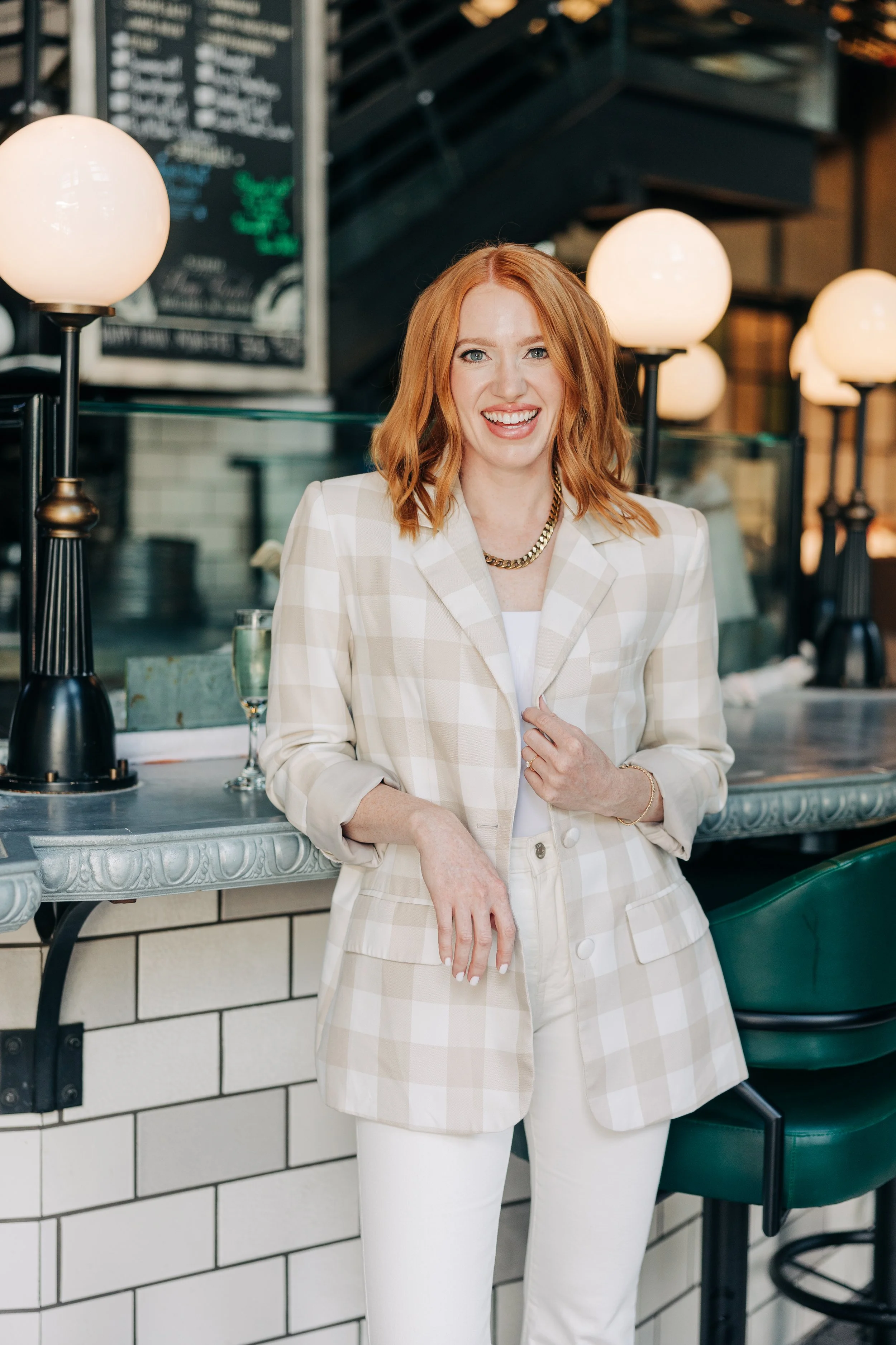 A woman with red hair smiling and wearing a cream-colored checkered blazer and white pants in a modern restaurant or cafe interior.