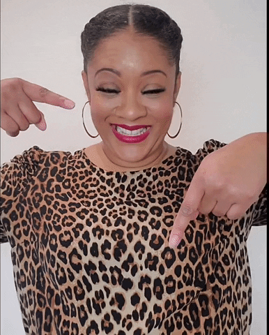 A woman wearing a leopard print top, smiling, with her eyes closed, pointing to her chest with both index fingers.
