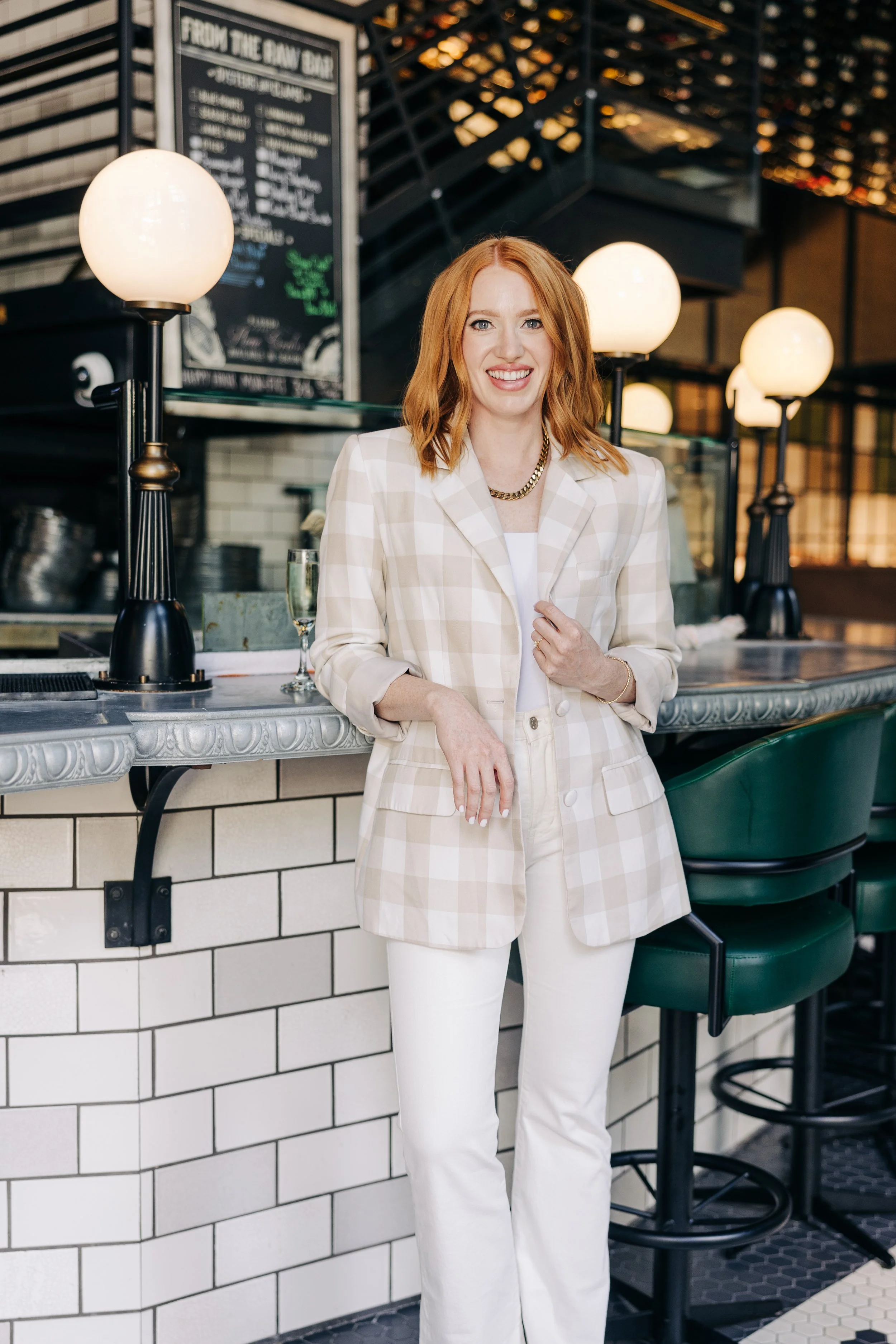A woman with red hair wearing a beige plaid blazer and white pants standing at a bar with a chalkboard menu in the background.