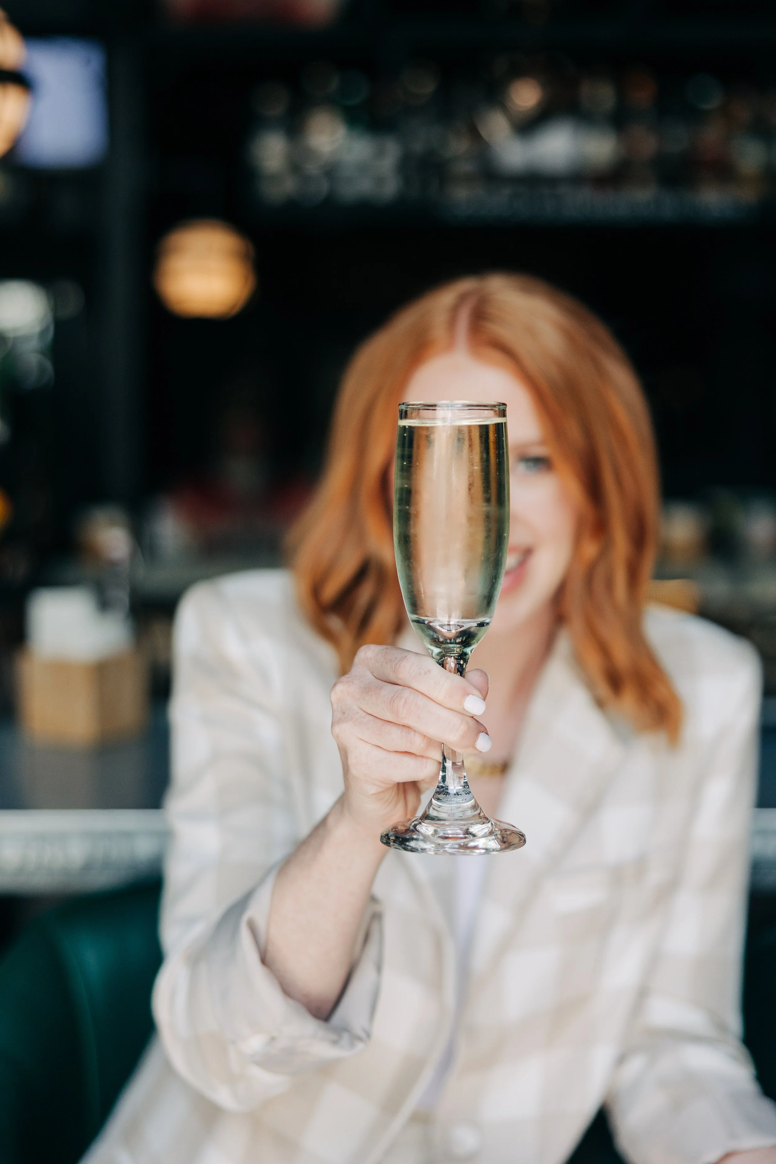 A woman with red hair smiling while holding up a glass of champagne in a restaurant or bar setting.