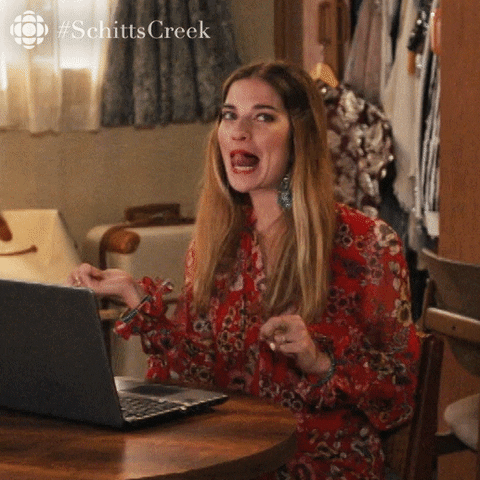 A woman with long hair, wearing a red floral dress, sitting at a table with a laptop, making a surprised or expressive face inside a room with hanging clothes and curtains.
