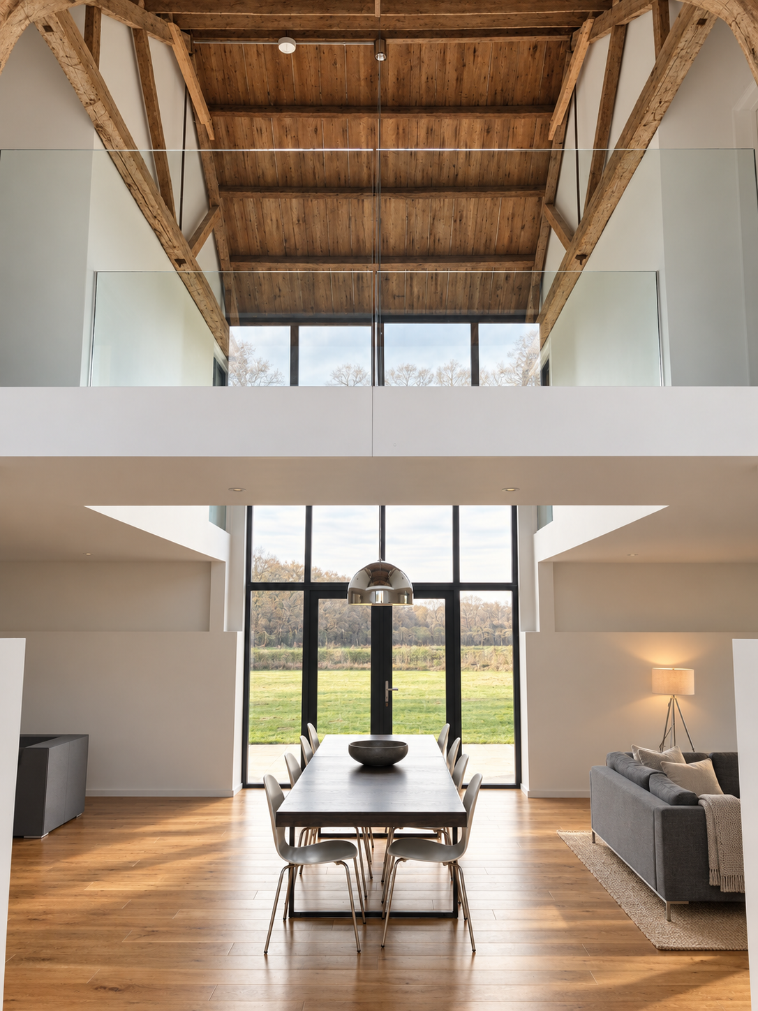 Double-height dining space under the original barn roof timbers, with a glass-balustraded gallery and full-height glazing at Little Nuntys