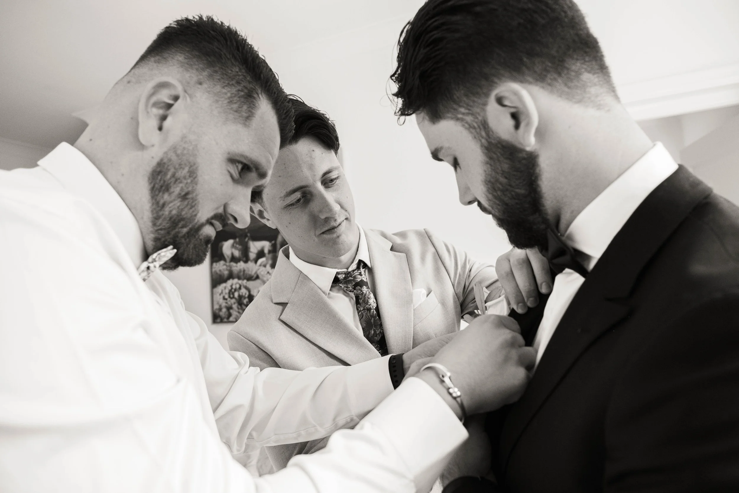 Trois hommes en costumes, dont deux avec une barbe, se préparant pour un événement formel, probablement un mariage ou une cérémonie, dans une pièce intérieure.