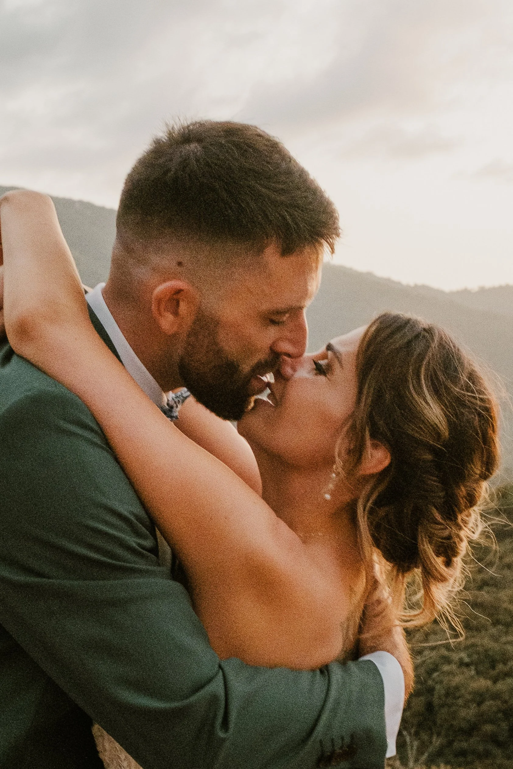 Un couple en tenue de mariage s'embrasse passionnément en plein air, avec un paysage de montagnes en arrière-plan pendant le coucher du soleil.