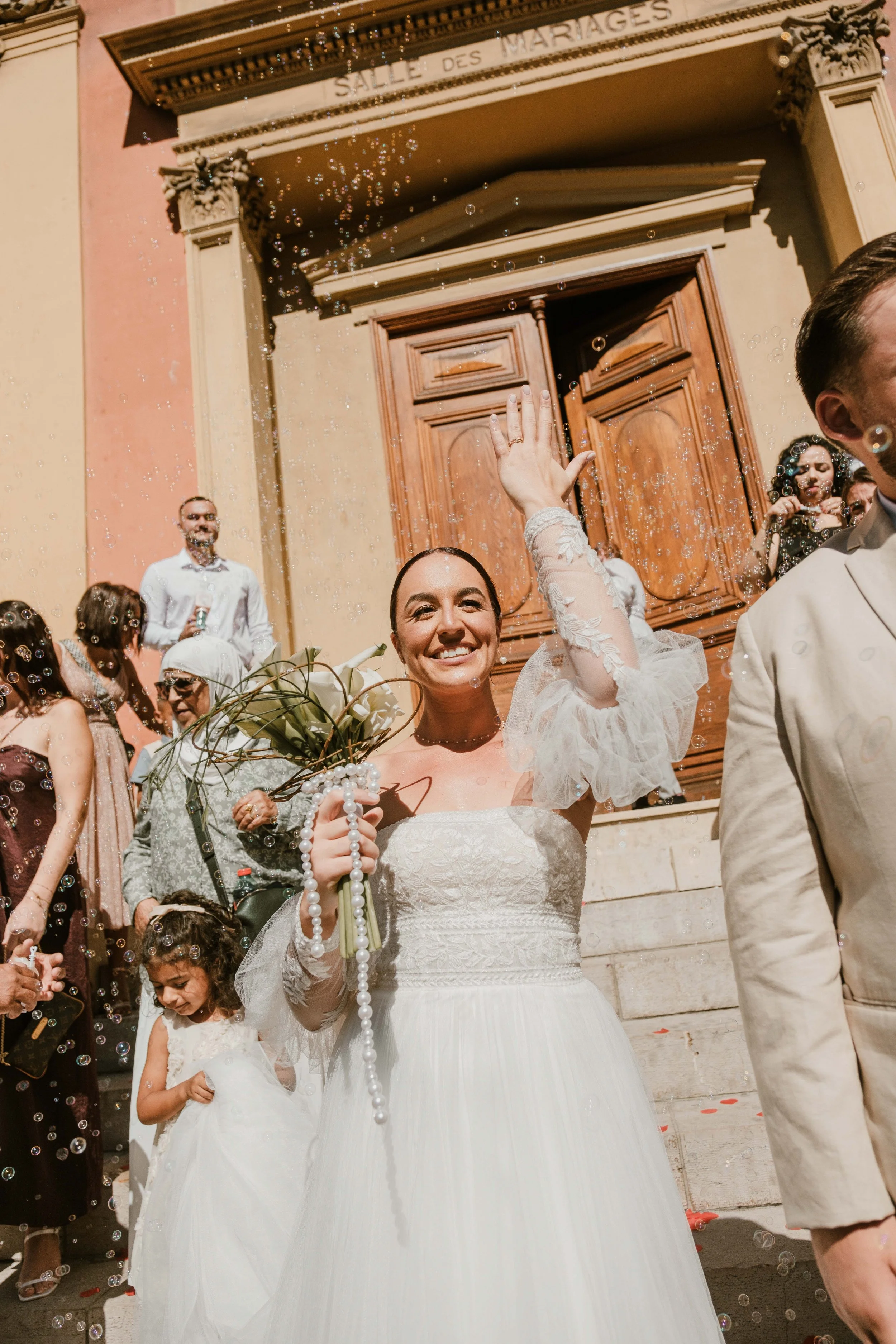Une mariée en robe blanche souriante, tenant un bouquet, fait signe de la main en descendant les marches d'une église lors d'un mariage, entourée d'invités et de bulles de savon