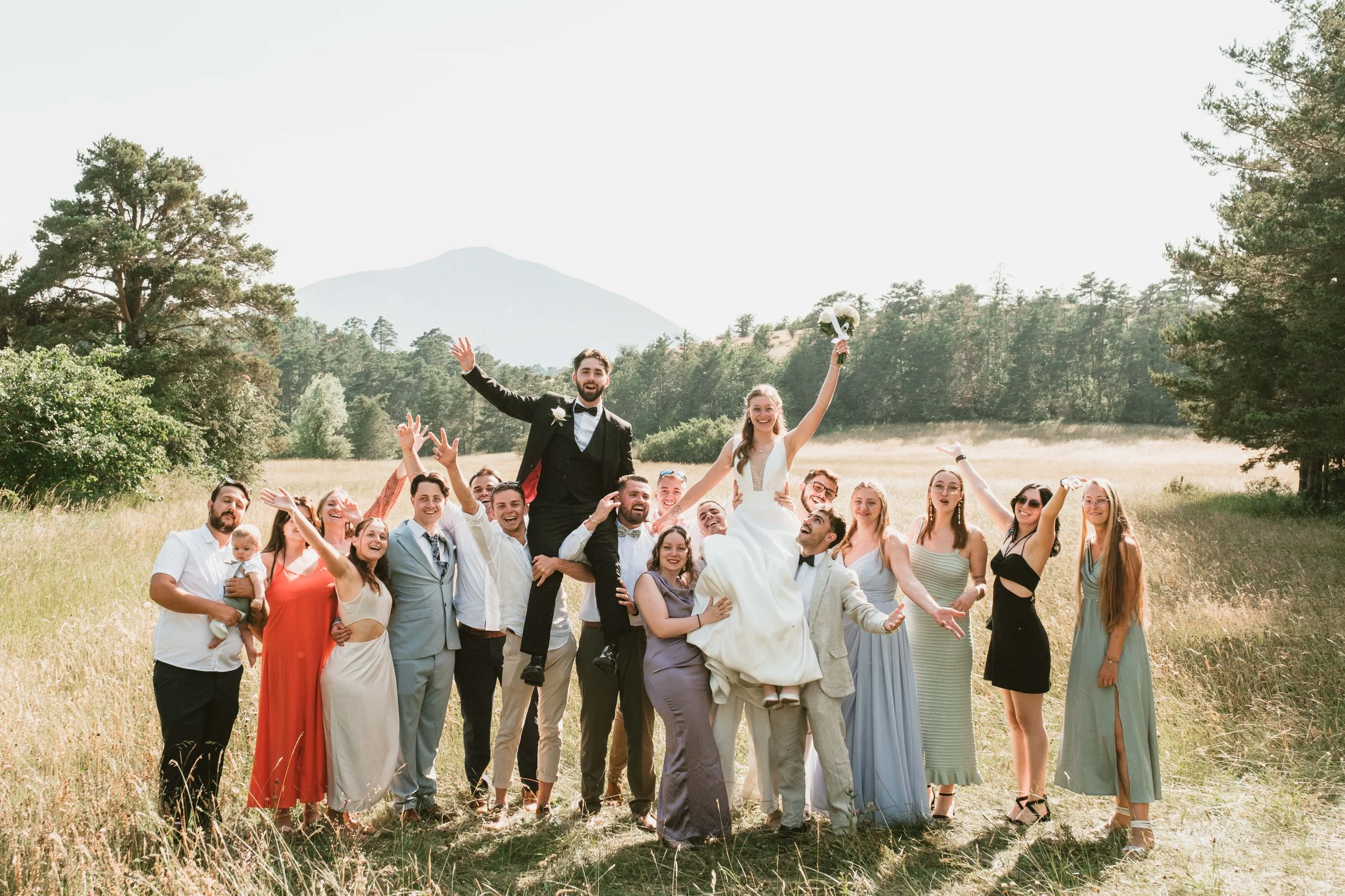 Groupe de personnes lors d'un mariage en plein air, avec un couple de mariés au centre, les mariés étant portés par des amis ou la famille, tous souriants et joyeux, dans un paysage naturel avec des arbres et des montagnes en arrière-plan.