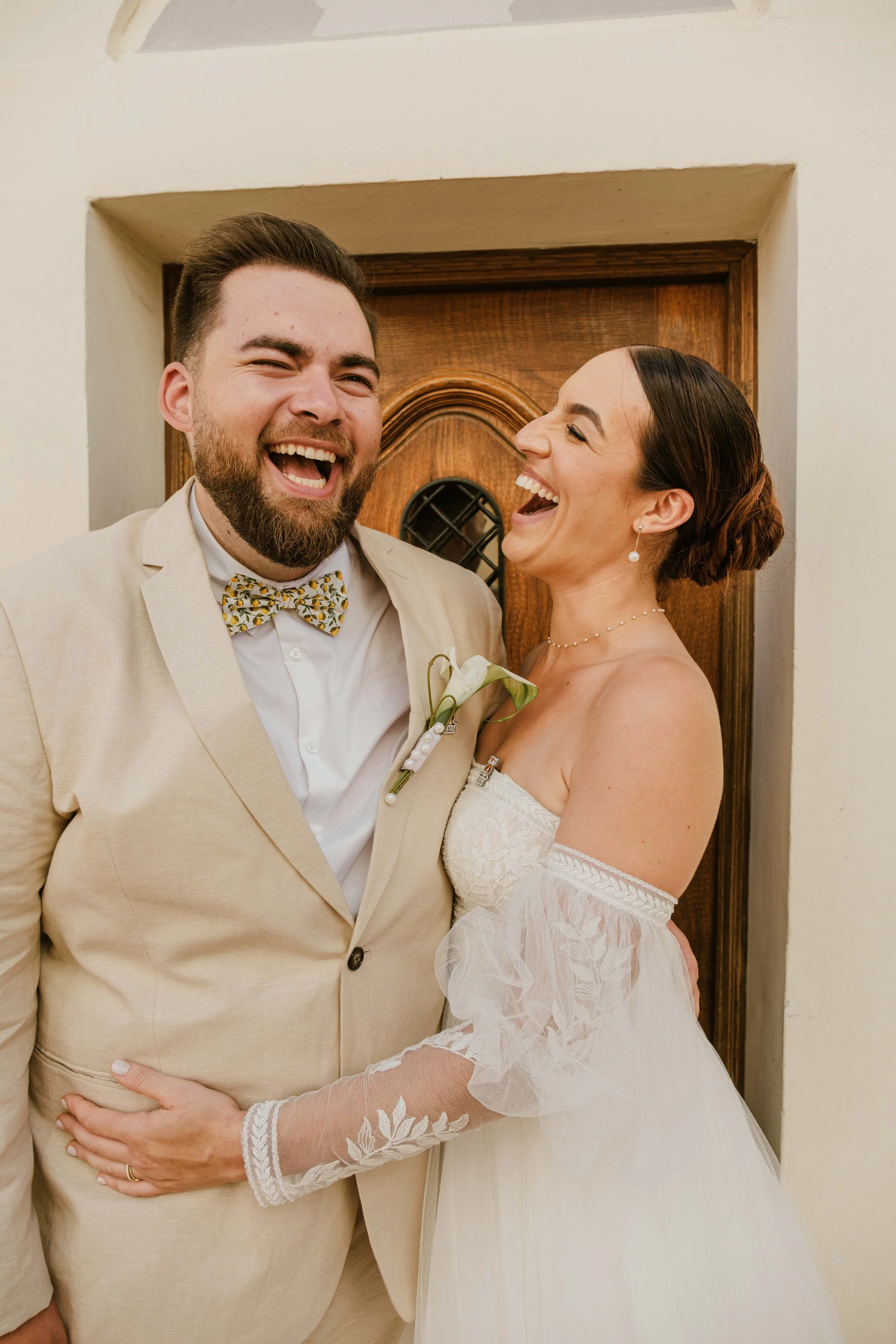 Un couple de mariés riant, debout devant une porte en bois, lors de leur mariage.