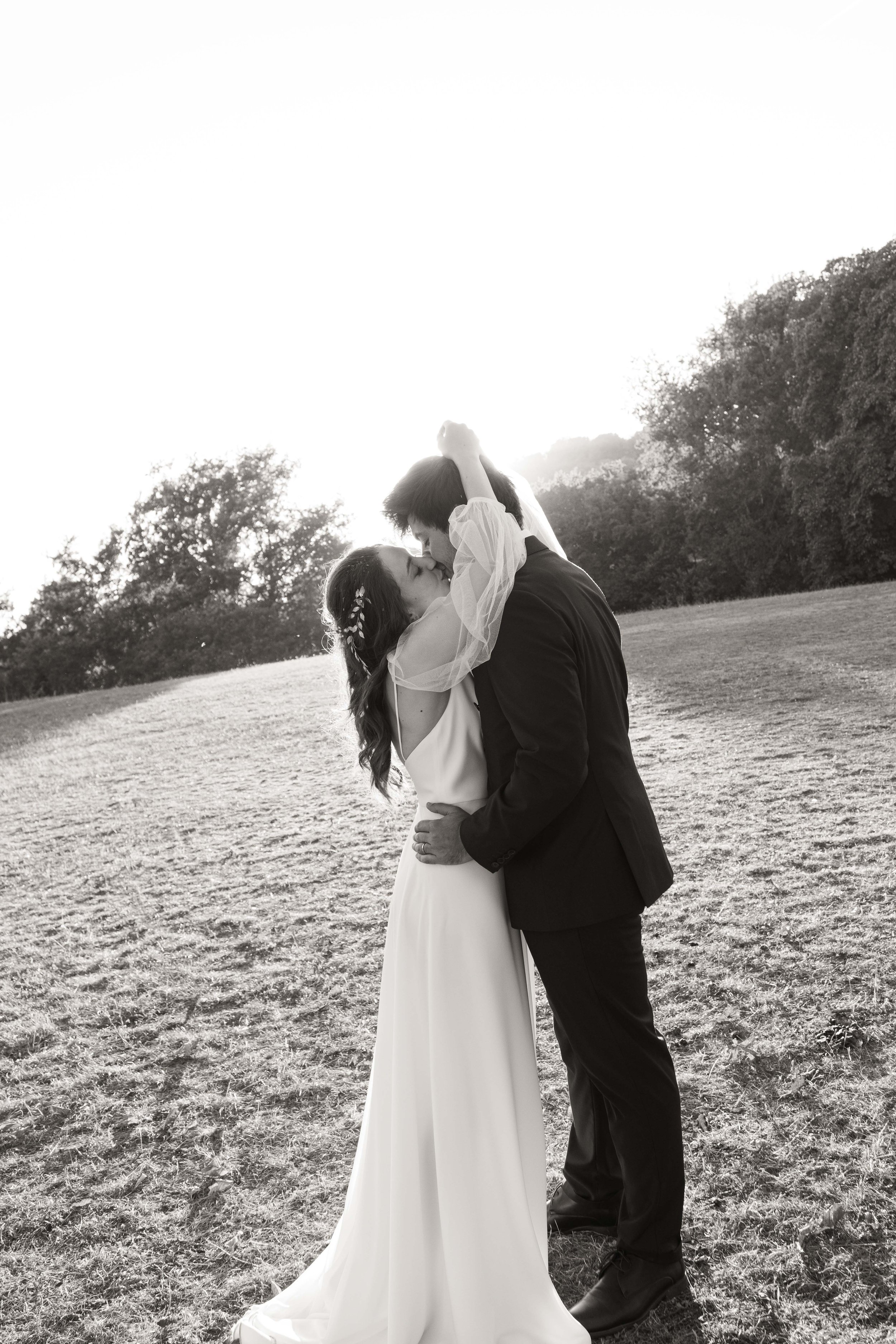 Un couple en mariage s'embrassant dans un champ en plein air, en noir et blanc, avec des arbres en arrière-plan et la lumière du soleil en arrière-plan.