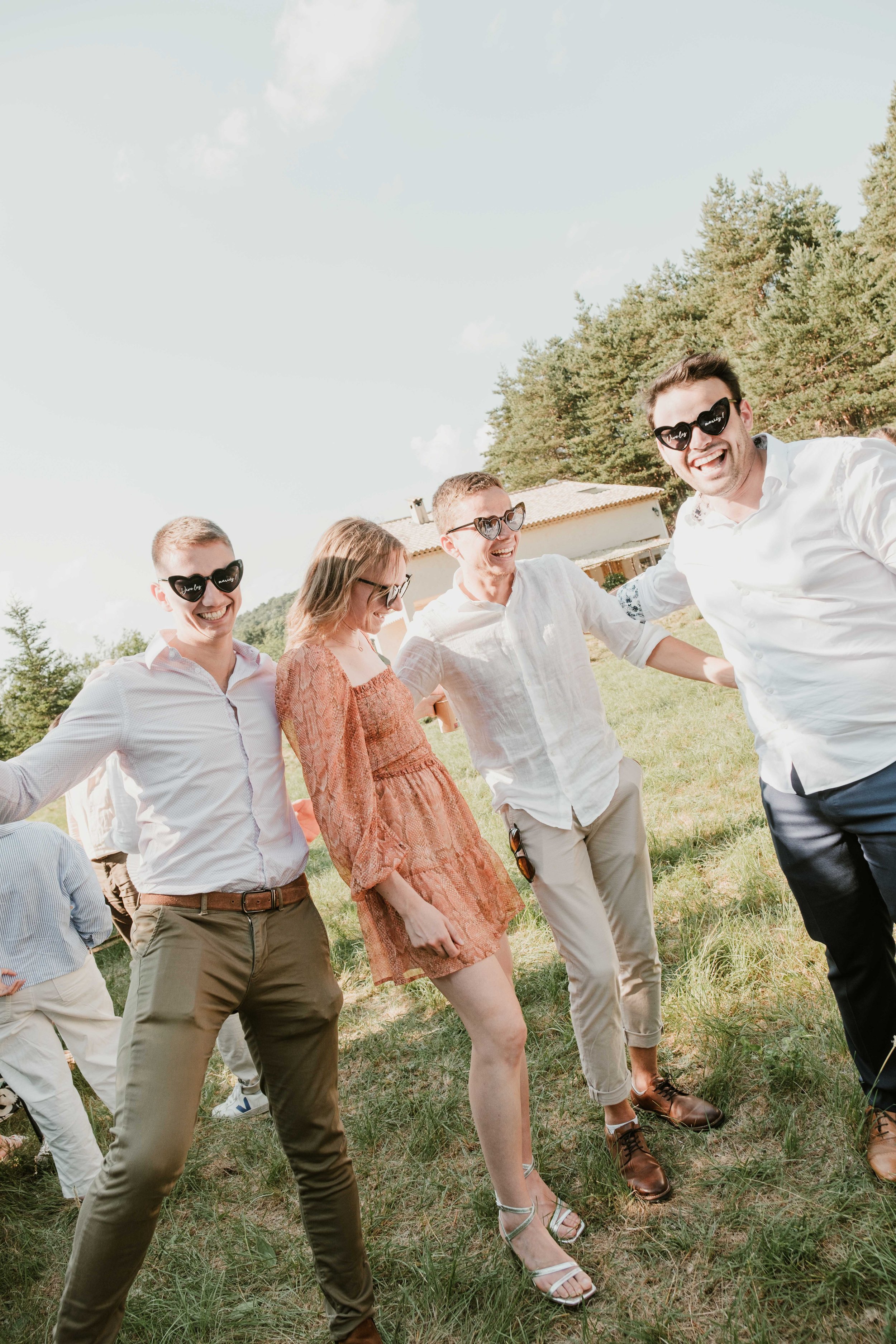 Groupe de cinq personnes souriantes, portant des lunettes de soleil en forme de cœur, dans un espace extérieur ensoleillé, avec des arbres et une maison en arrière-plan.