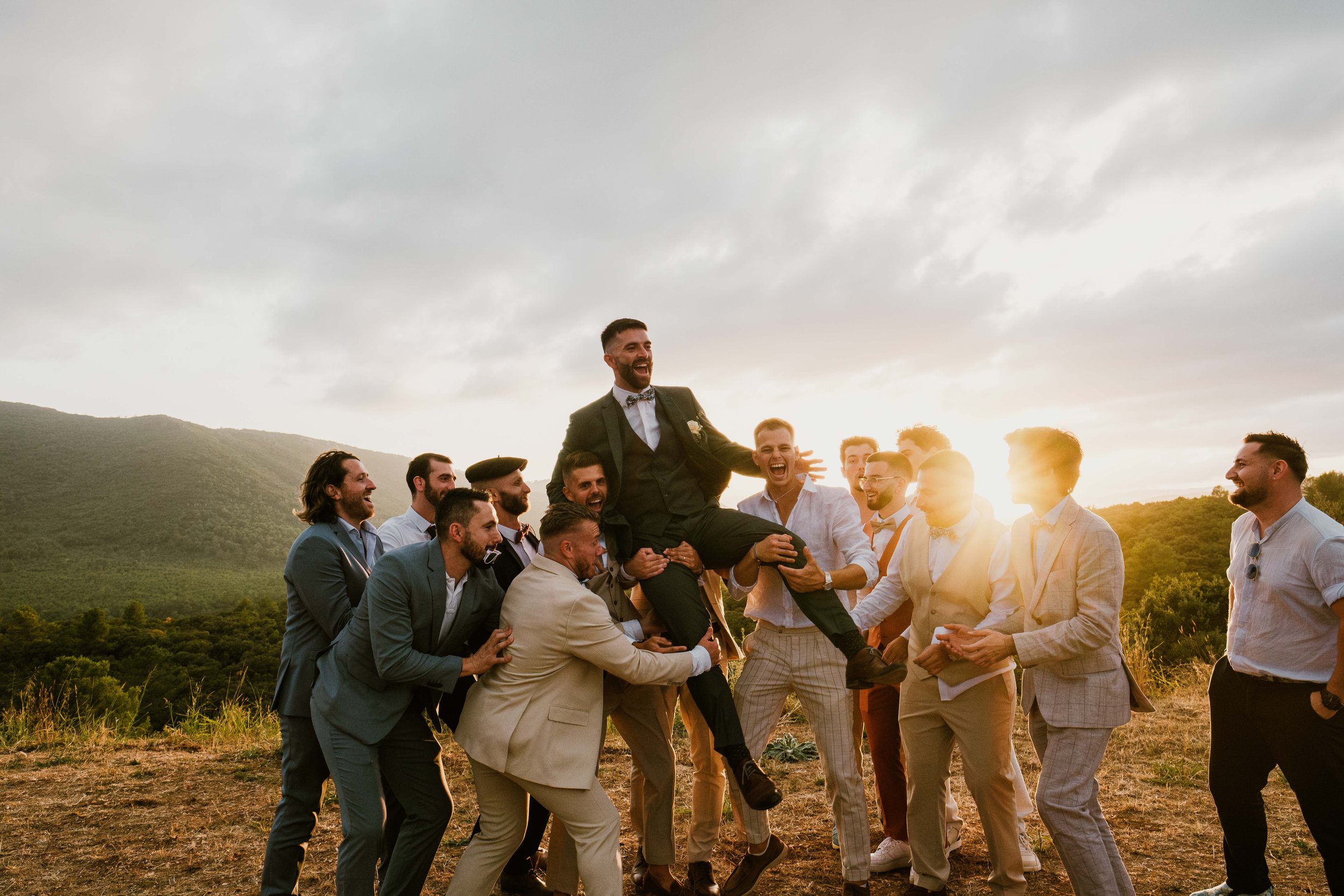 Un groupe d'hommes habillés en costumes, célébrant lors d'une occasion spéciale en plein air au coucher du soleil, avec des montagnes en arrière-plan.