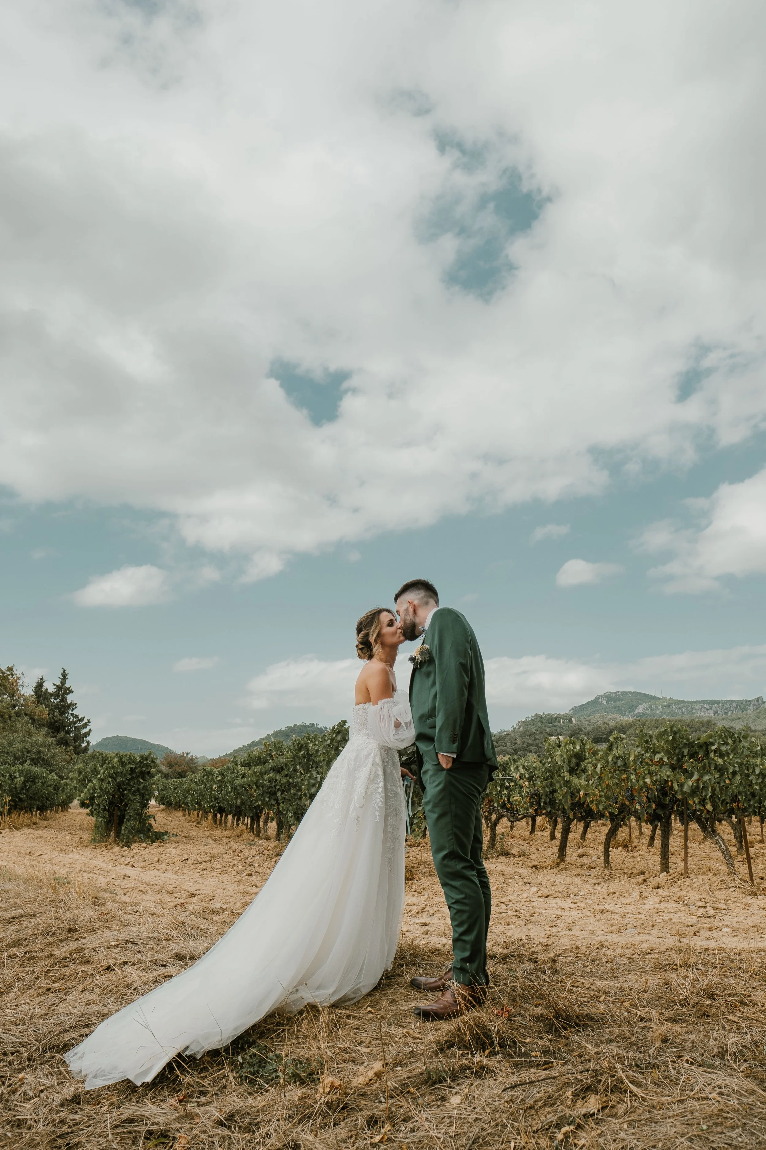 Un couple marié, la femme en robe de mariée blanche et le homme en costume vert, s'embrassant dans un vignoble avec un ciel nuageux.