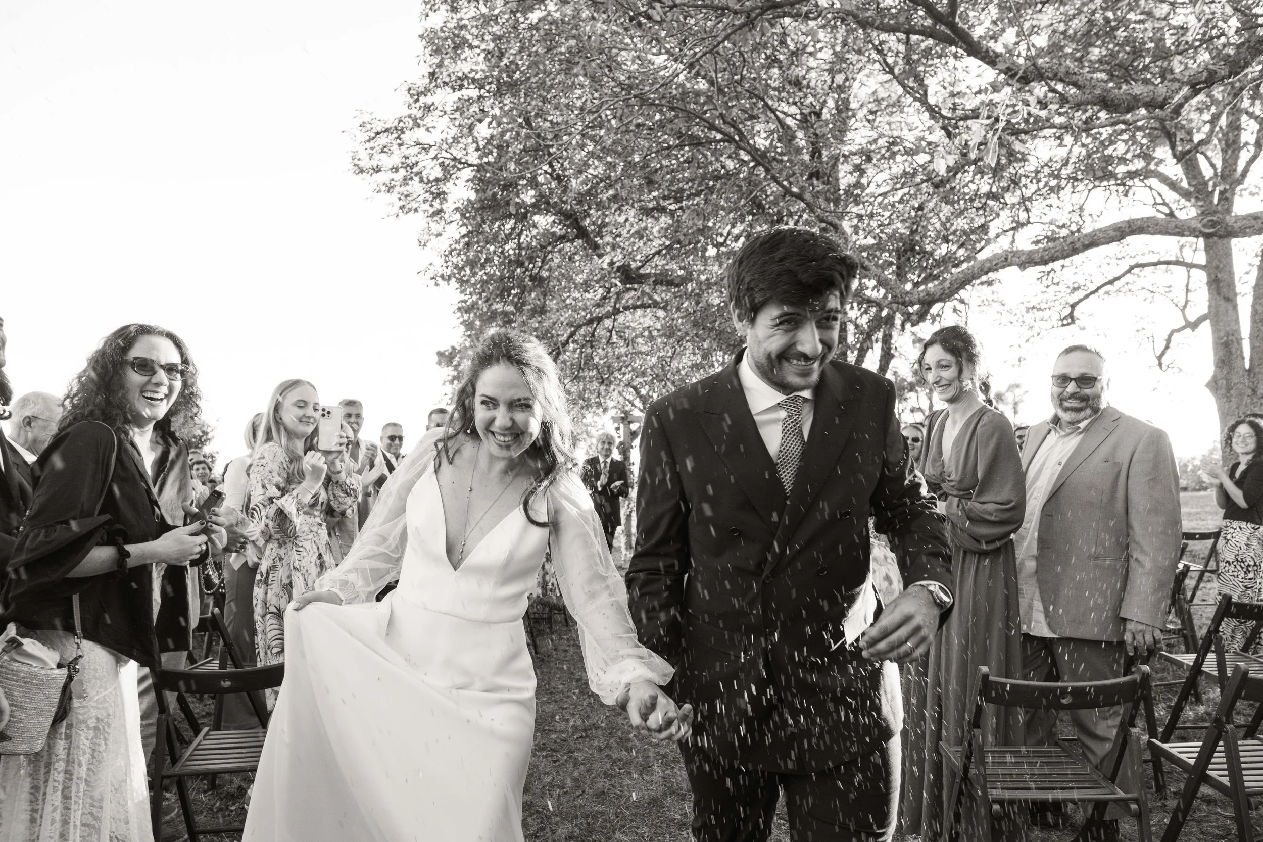 Un couple de mariés souriants marche main dans la main lors de leur mariage en plein air, entourés d'invités qui prennent des photos et applaudissent, sous de grands arbres.