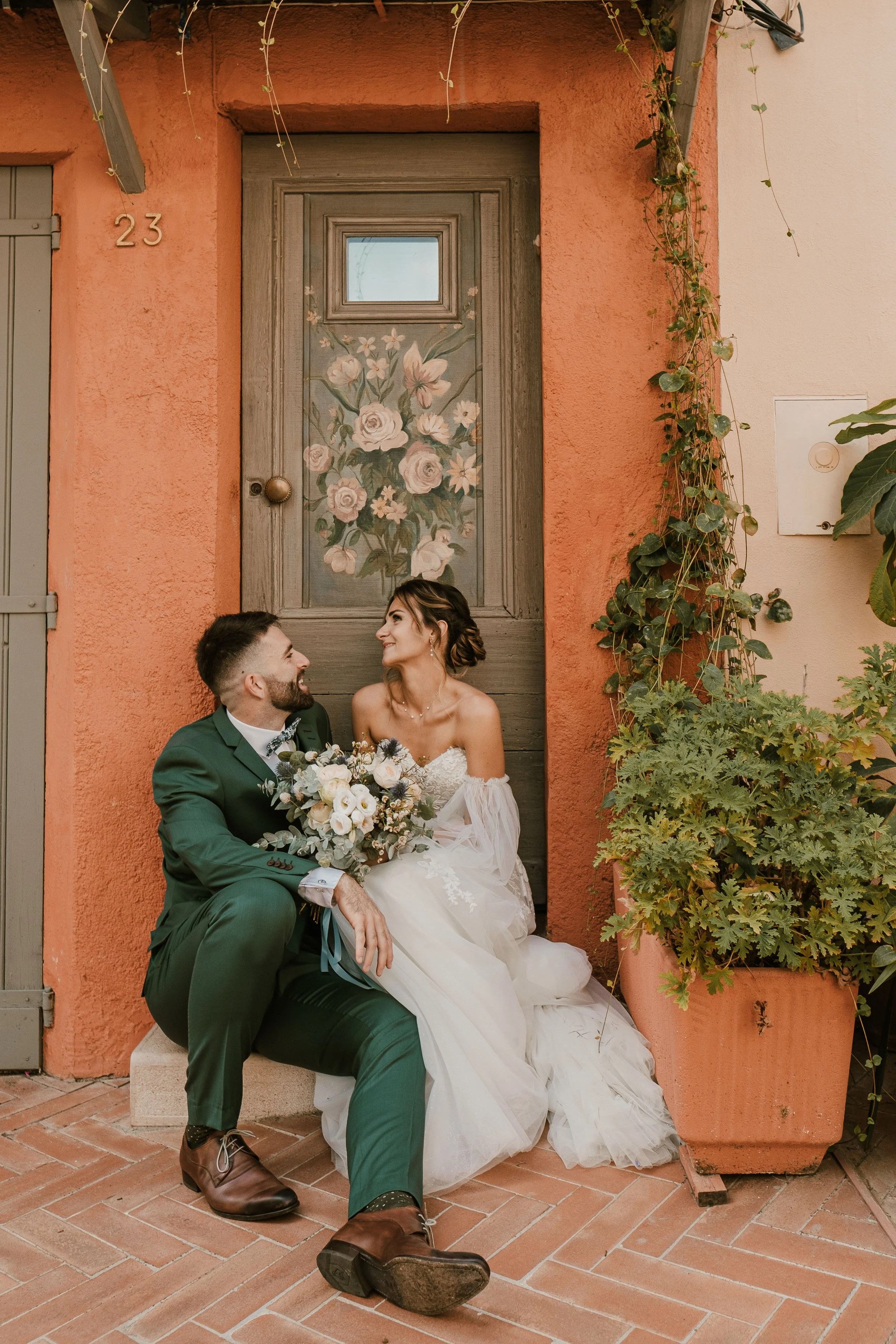 Un couple marié assis devant une porte en bois avec une peinture de fleurs, entouré de plantes en pot, dans un décor coloré à Mouans-Sartoux.