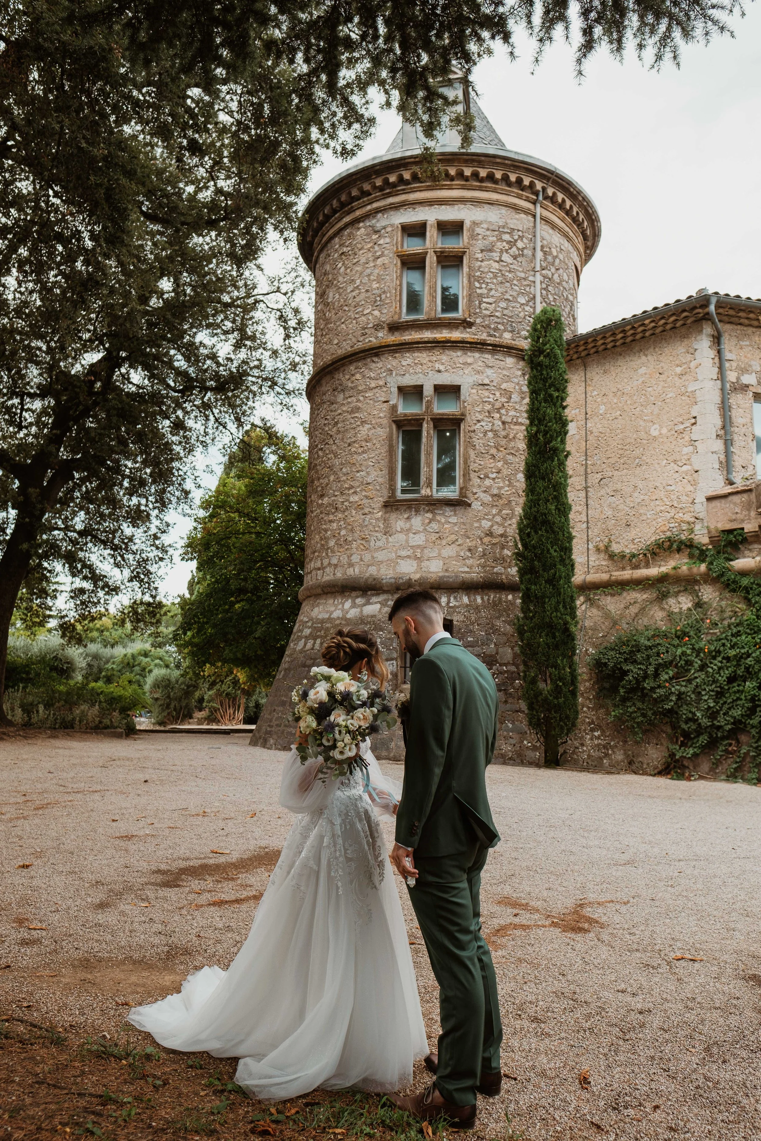 Un couple en robe de mariée et costume de mariage, le couple se tenant la main devant un vieux bâtiment en pierre avec une tour cylindrique, entourés d'arbres et de végétation.