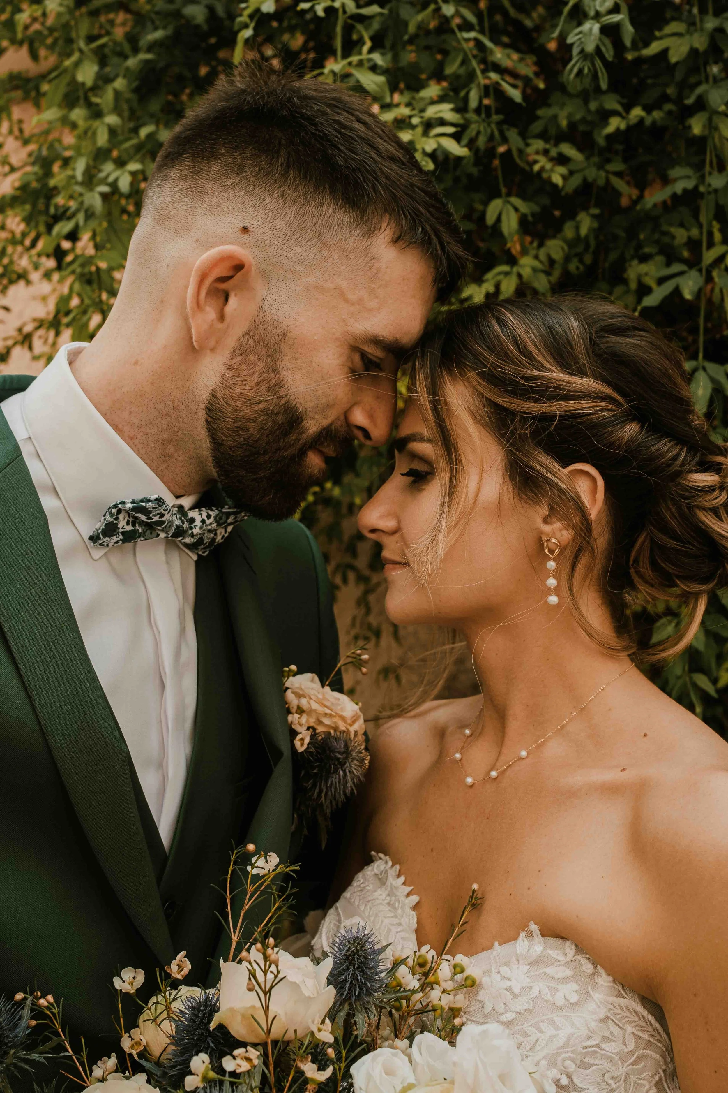Un couple de mariage en gros plan, face à face, avec la tête touchant, tenant un bouquet de fleurs, devant un arrière-plan de feuillage.