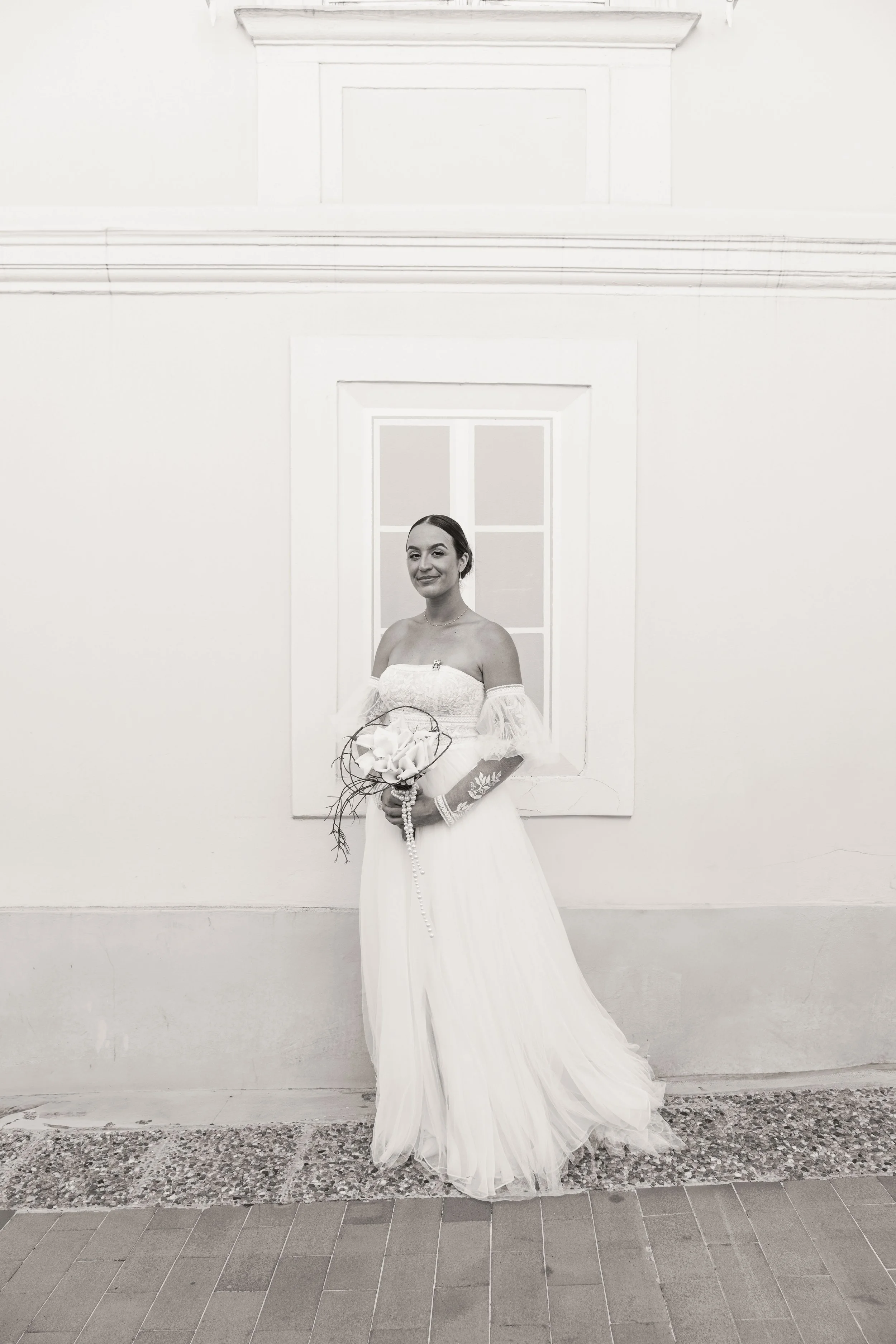 Une femme en robe de mariage tient un bouquet de fleurs devant une fenêtre, photo en noir et blanc.