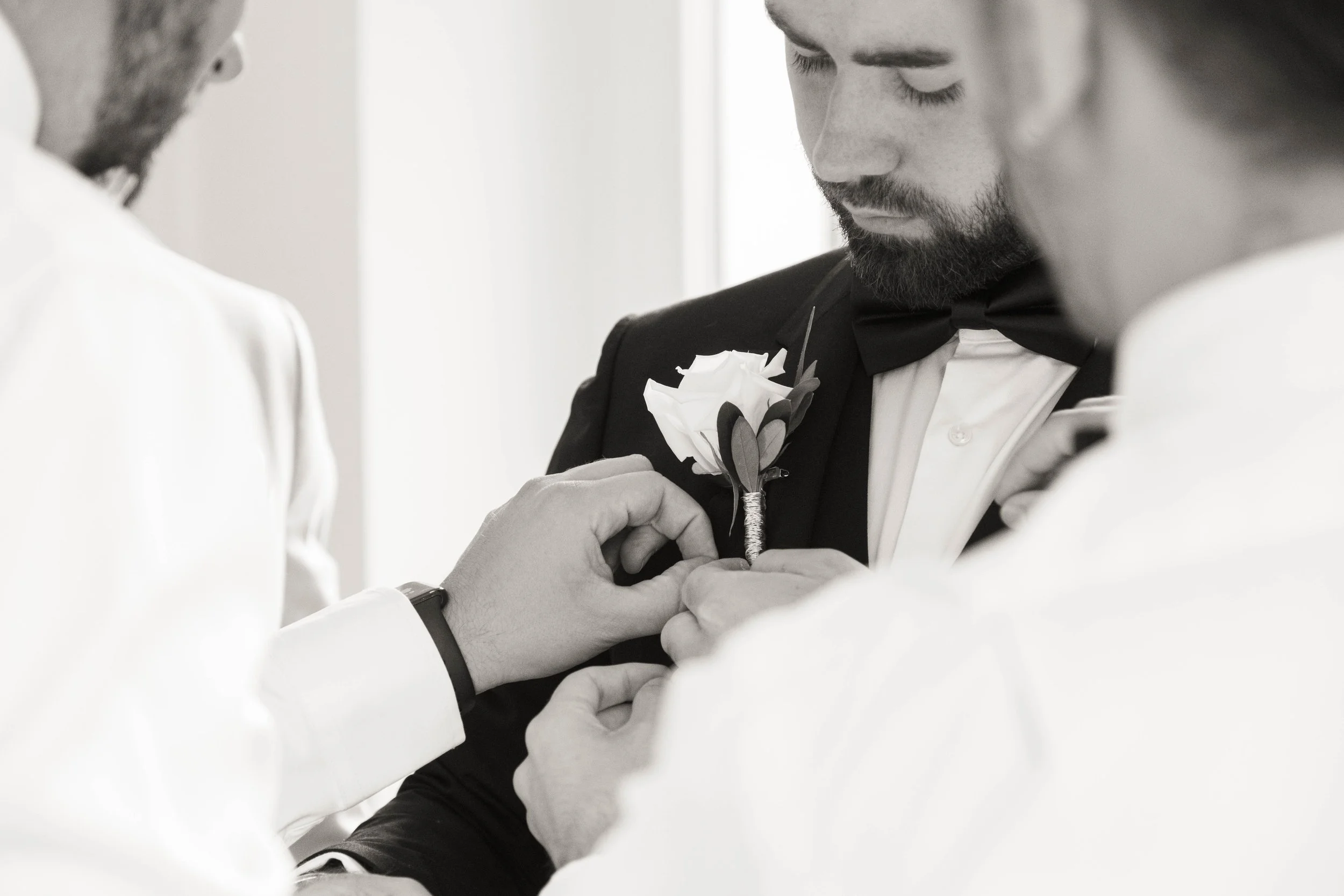 Groom helping best man pin a boutonnière on his tuxedo during wedding preparations.