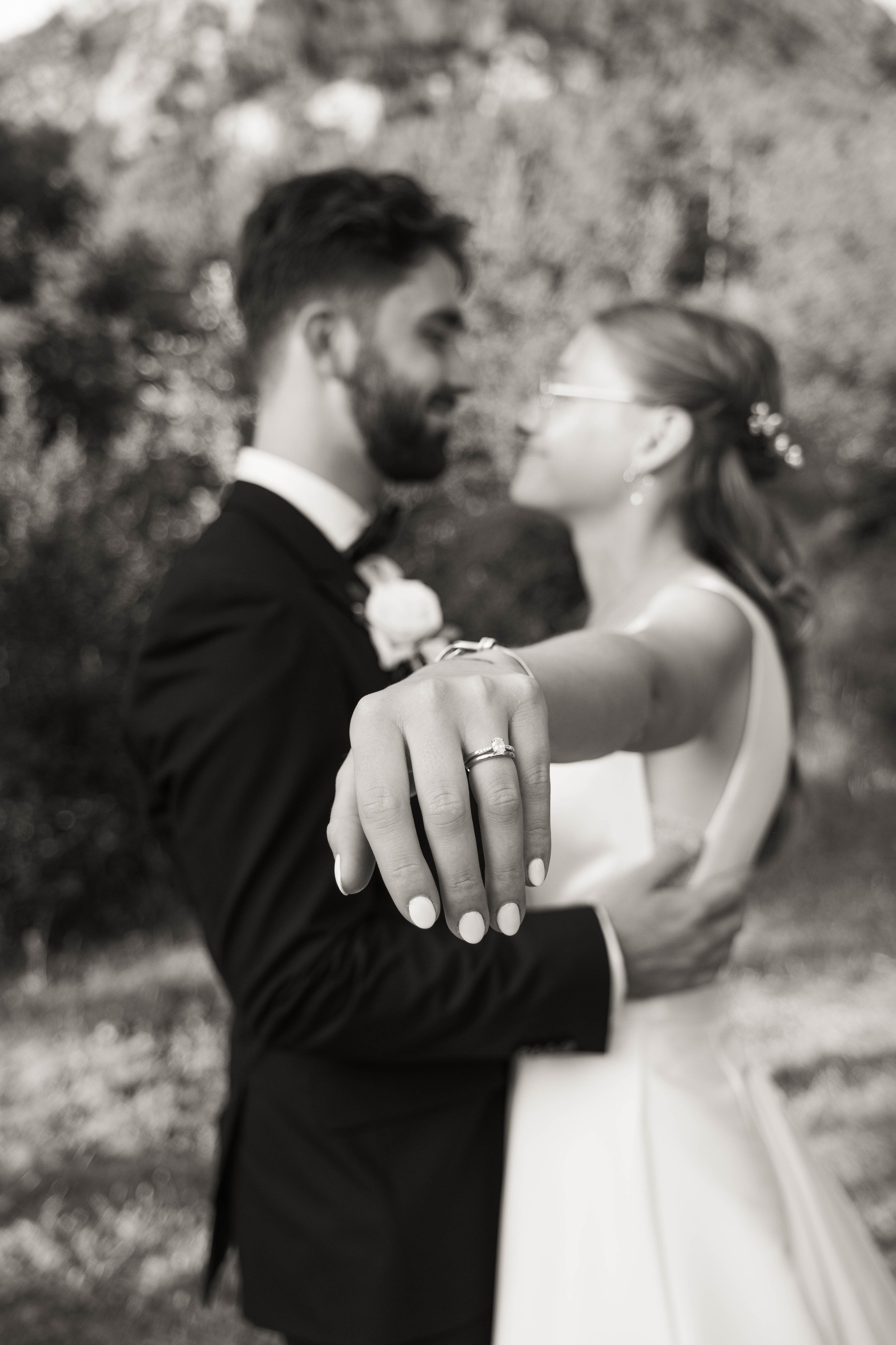 Un couple de mariés en noir et blanc, l'homme en costume et la femme en robe de mariage, se tenant de face et se regardant, avec la main de la femme montrant leur alliance