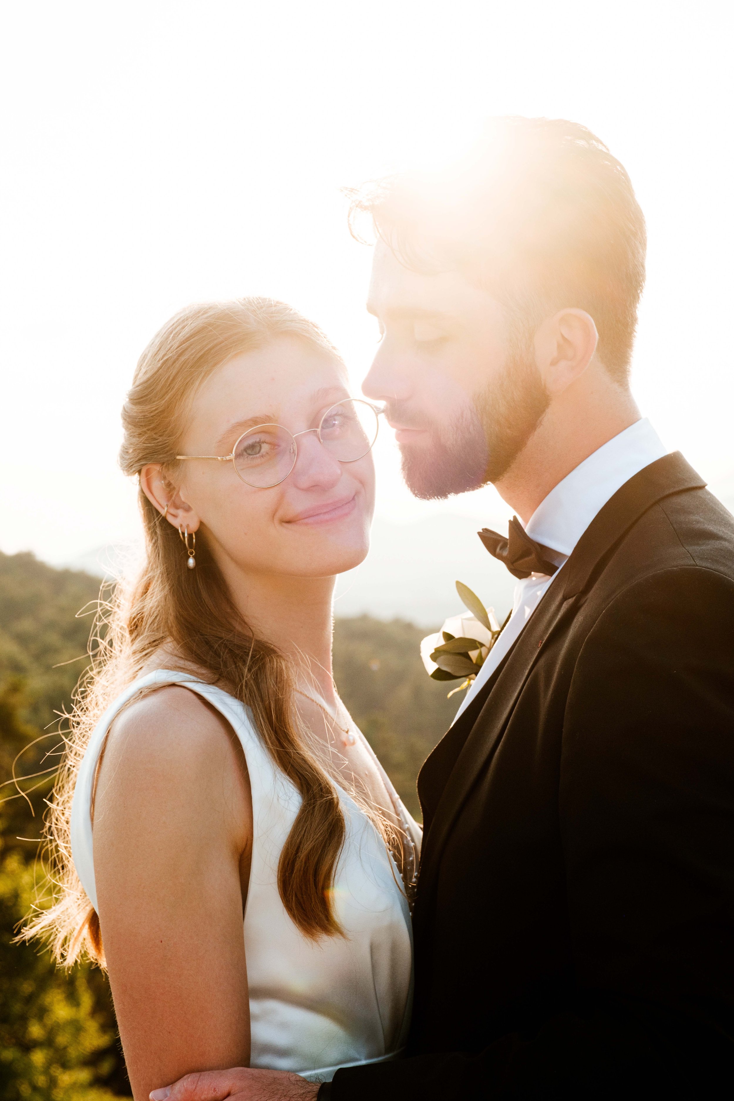 Un couple habillé en mariage, la femme porte des lunettes et une robe blanche, l'homme porte un costume noir avec une boutonnière, le soleil brille derrière eux, en plein air.