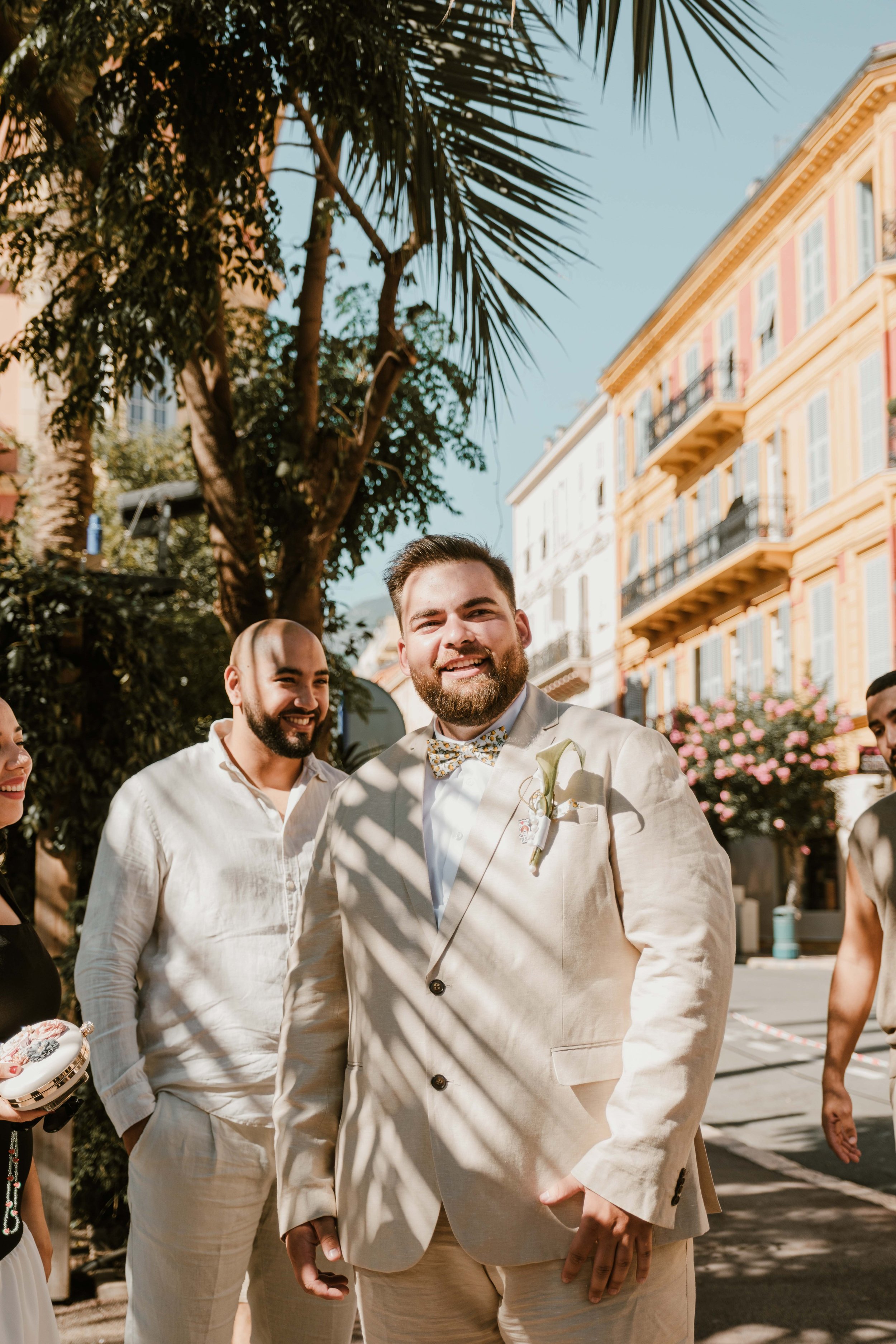 Groupe de personnes souriantes lors d'un mariage en plein air, avec un homme en costume beige avec une boutonnière, sous un palmier