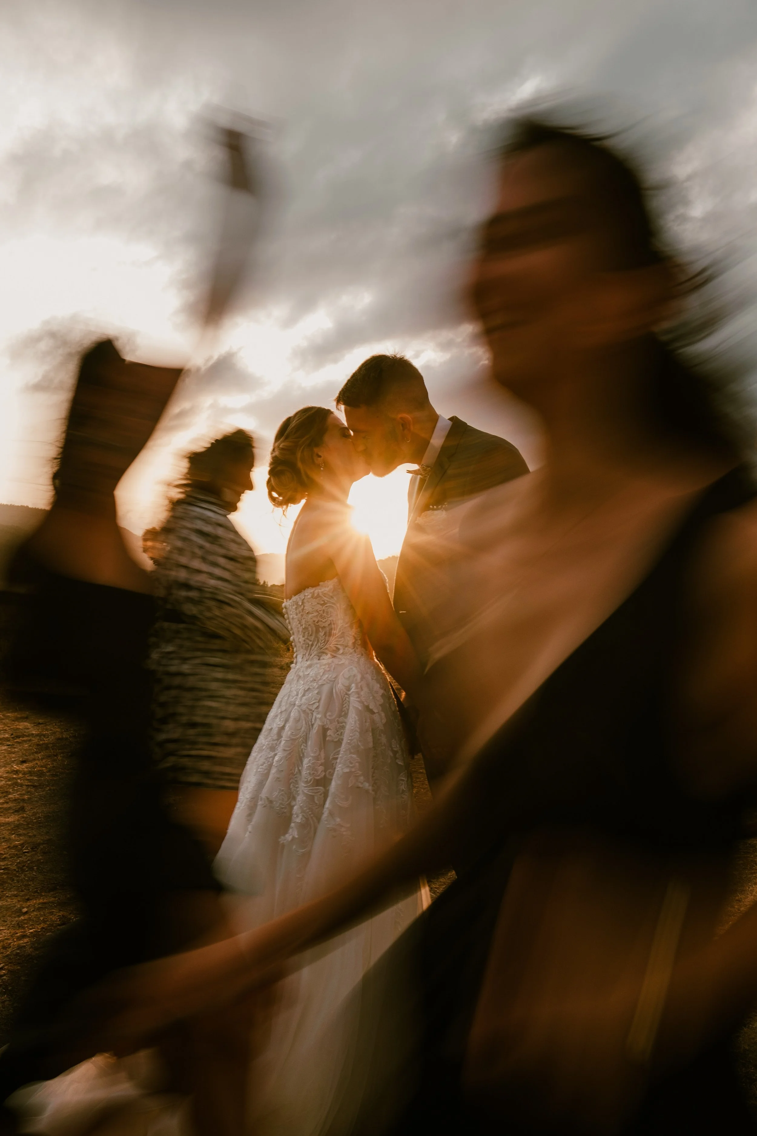 Un couple de mariés s'embrassant lors de leur mariage au coucher du soleil, entourés d'autres personnes floues en mouvement.
