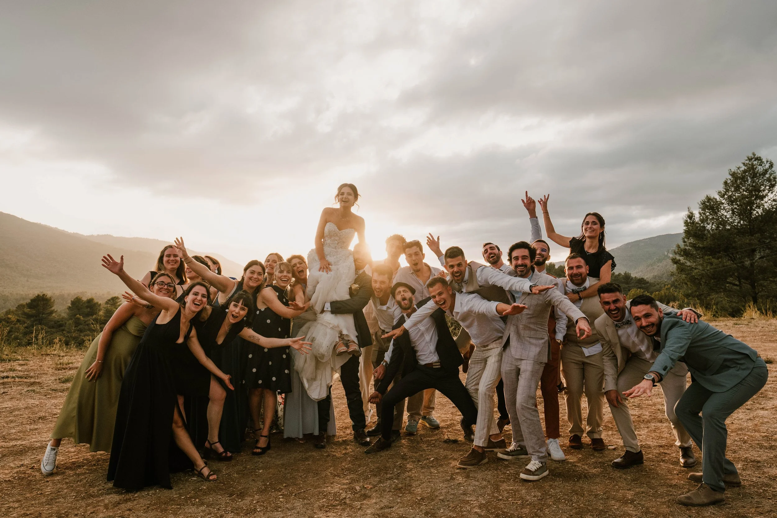 Un groupe de personnes en tenue de mariage posant en plein air avec une mariée sur les épaules d'une personne, sous un ciel nuageux, dans un paysage naturel avec des montagnes et des arbres.