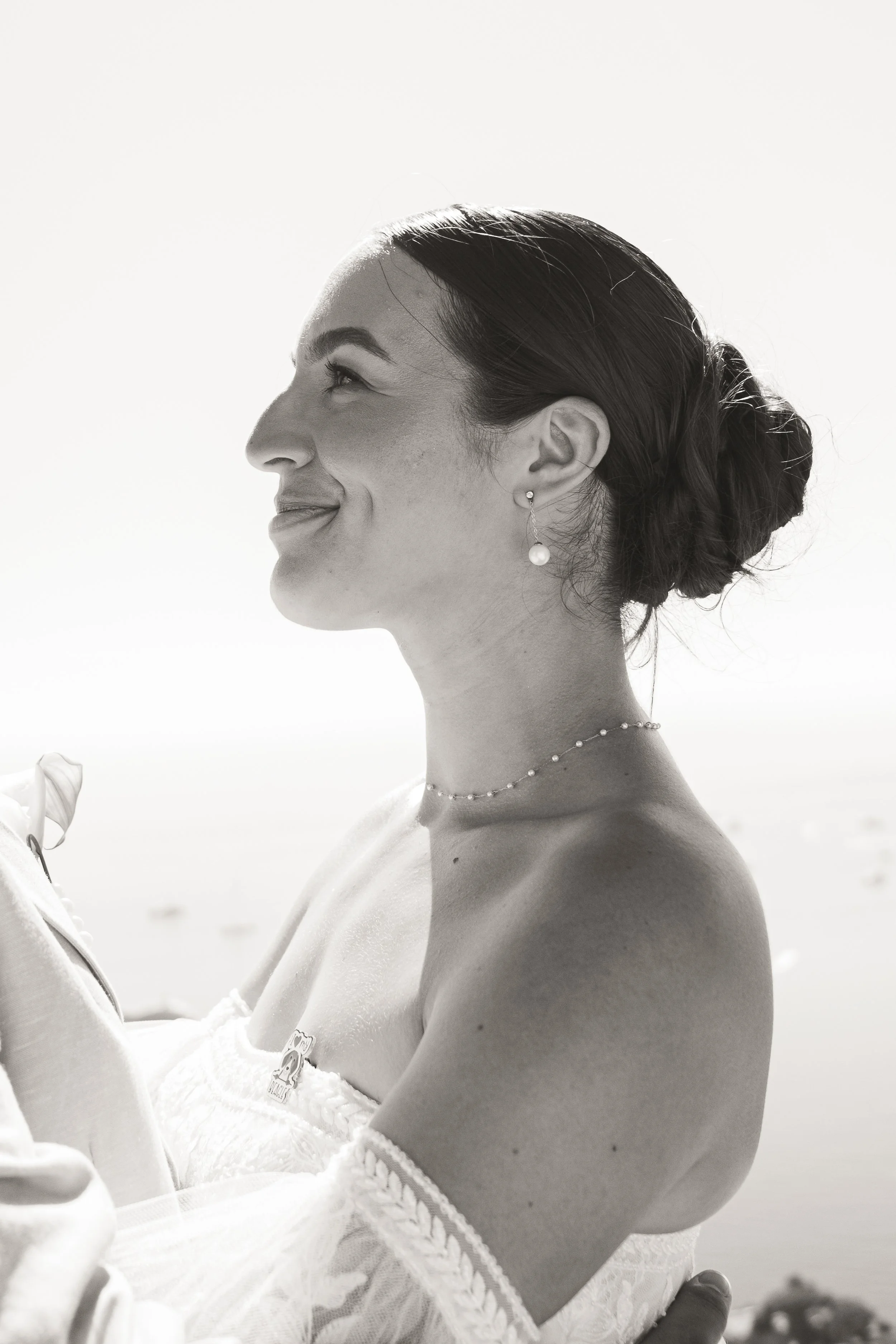 Portrait en noir et blanc d'une femme portant des bijoux, regardant de profil, avec une coiffure en chignon, devant un fond lumineux, probablement à la plage.