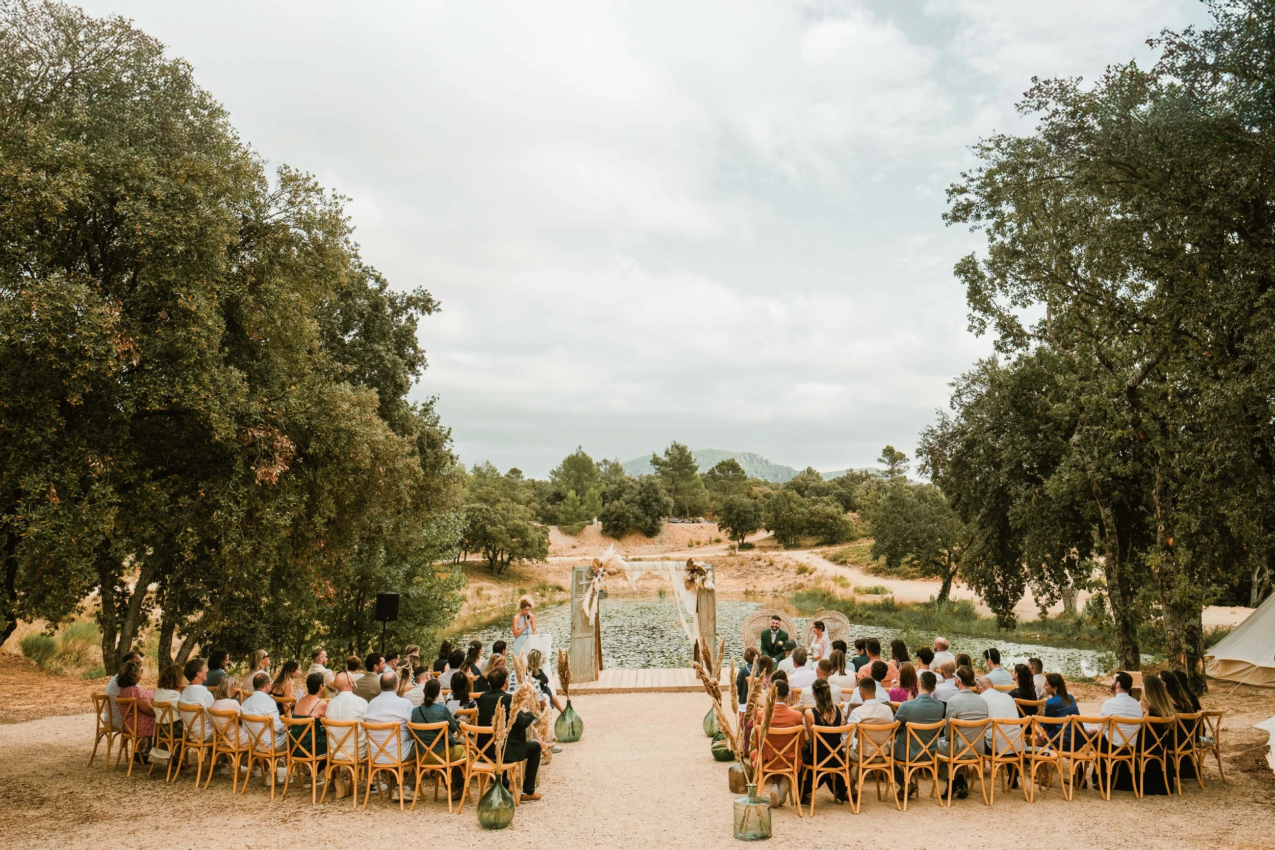 Cérémonie de mariage en plein air devant un lac, avec un arc décoré au centre et des invités assis sur des chaises en bois, entourée d'arbres et d'une nature paisible.