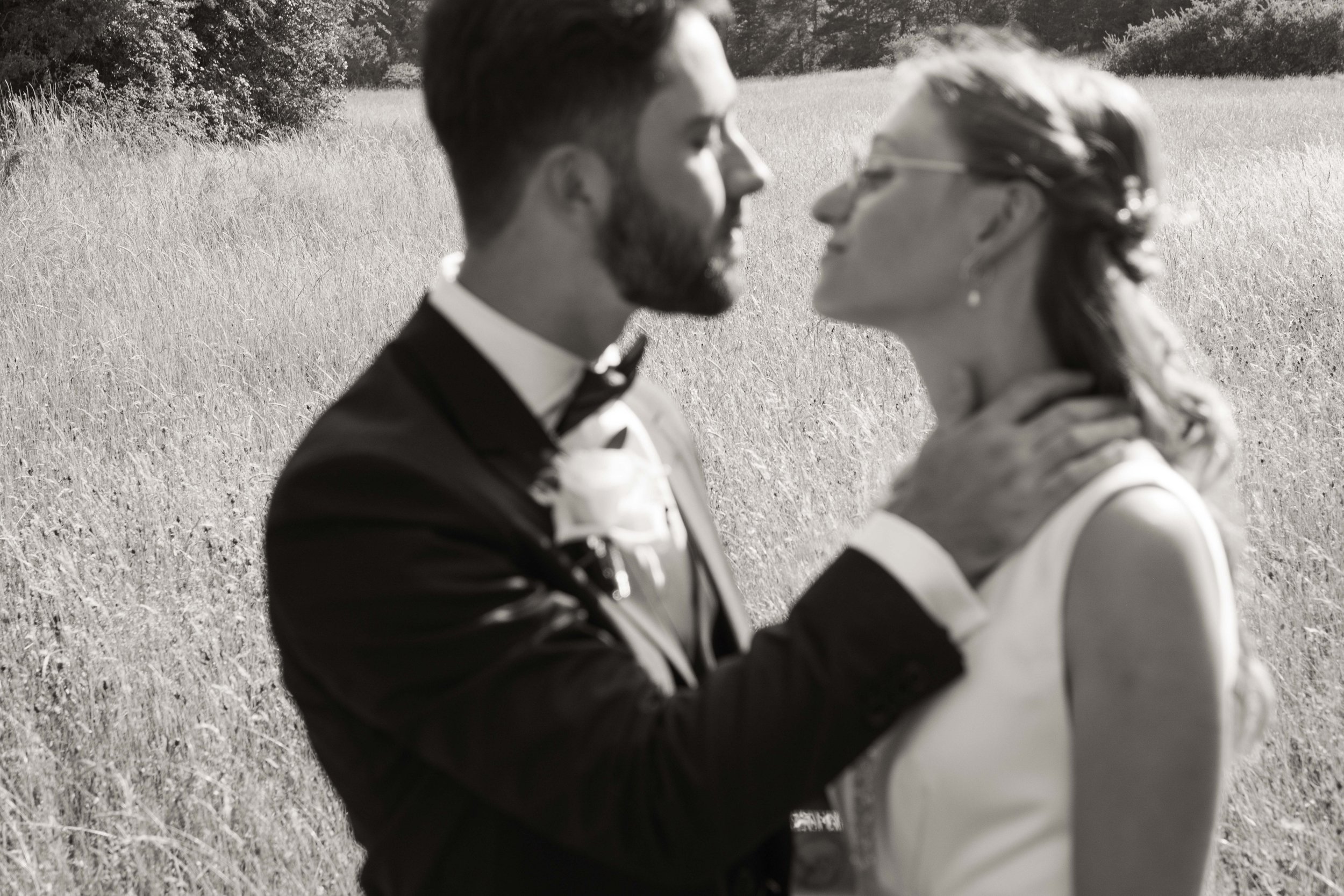 Un couple en mariage en plein air, l'homme en costume noir et la femme en robe blanche, se regardent tendrement dans un champ verdoyant.
