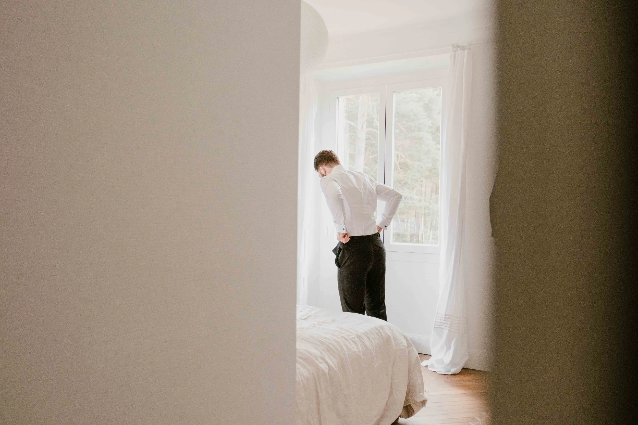 Un homme habillé en chemise blanche et pantalon noir regarde par une fenêtre dans une chambre lumineuse.