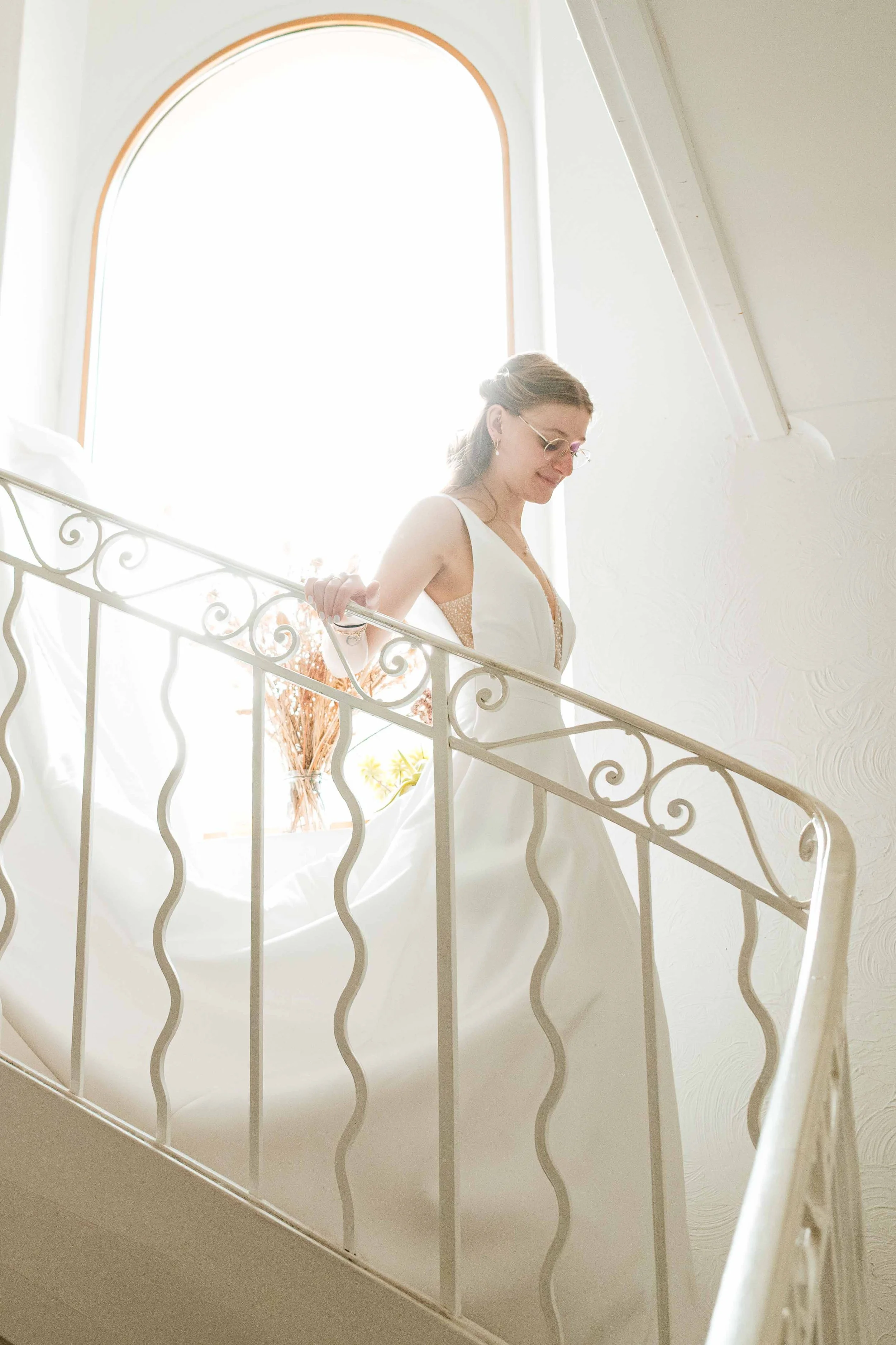 Une femme en robe de mariée descend un escalier en métal blanc, éclairée par une grande fenêtre arqué, avec un bouquet derrière elle.