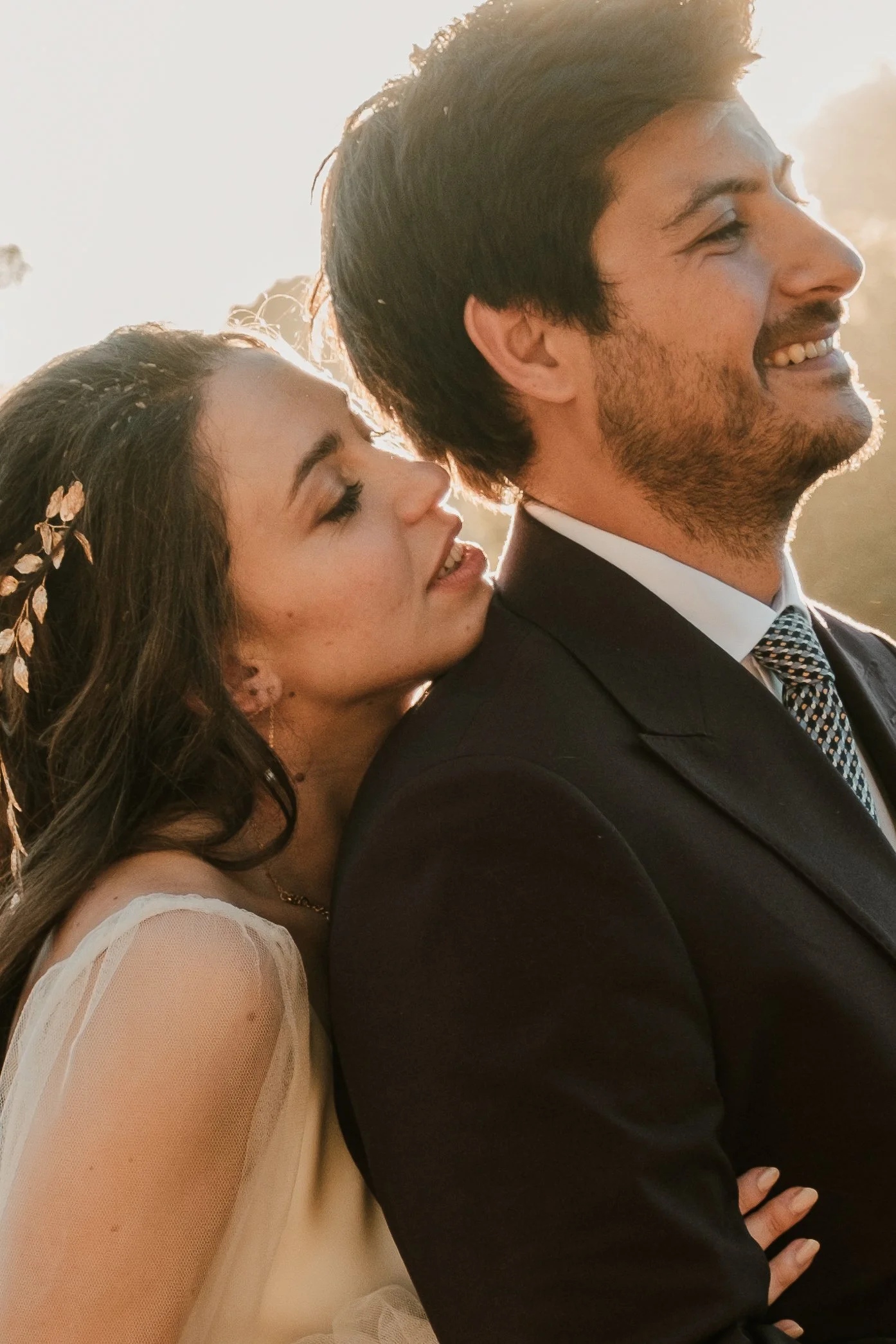 Un couple habillé pour un mariage, l'homme en costume et la femme en robe blanche, posant de face avec le soleil couchant en arrière-plan, souriant en intimité.