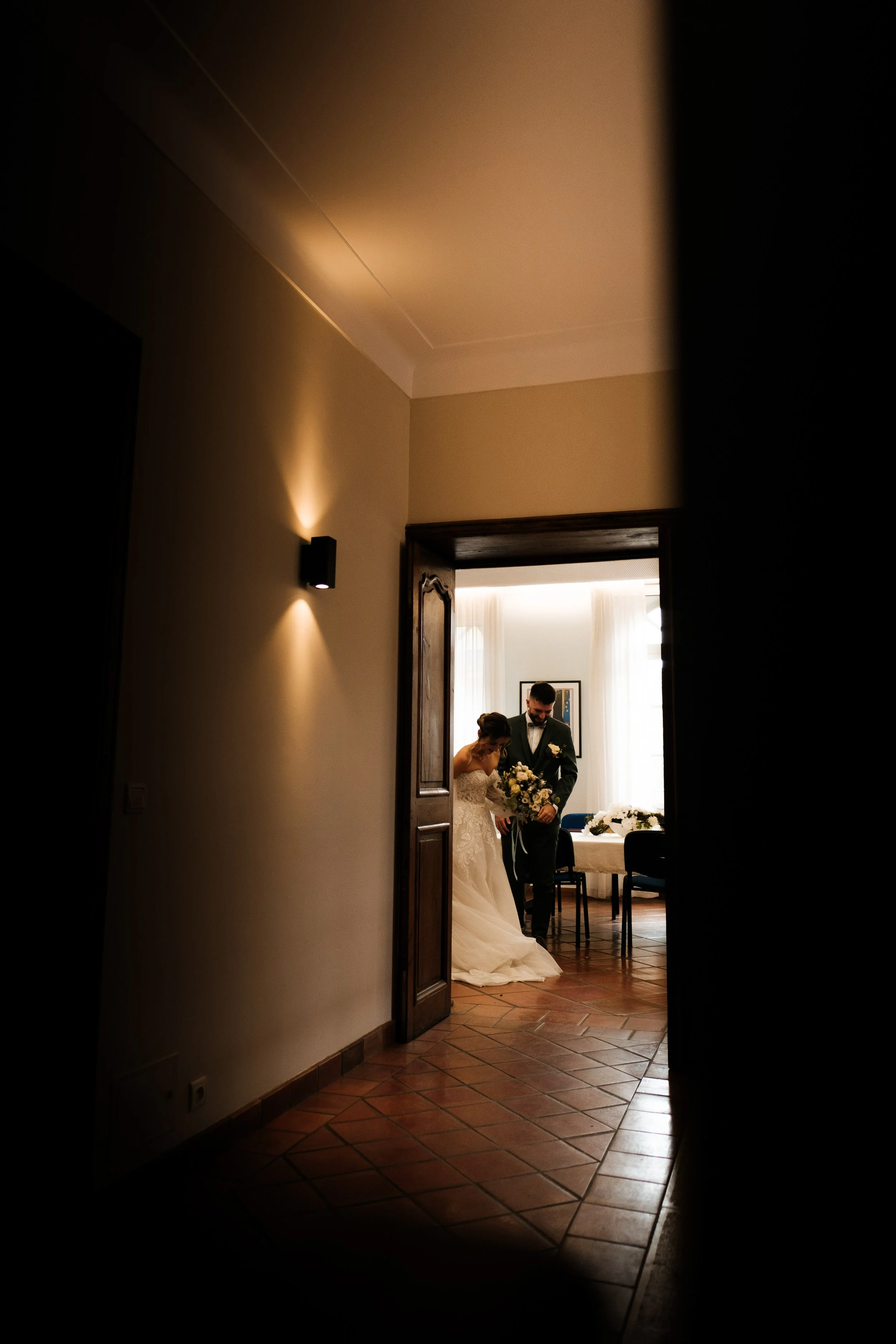 Jeune couple marié dans une pièce lumineuse, vue à travers une porte partiellement ouverte, avec des murs clairs, un sol en carrelage, et une table avec des fleurs en arrière-plan.