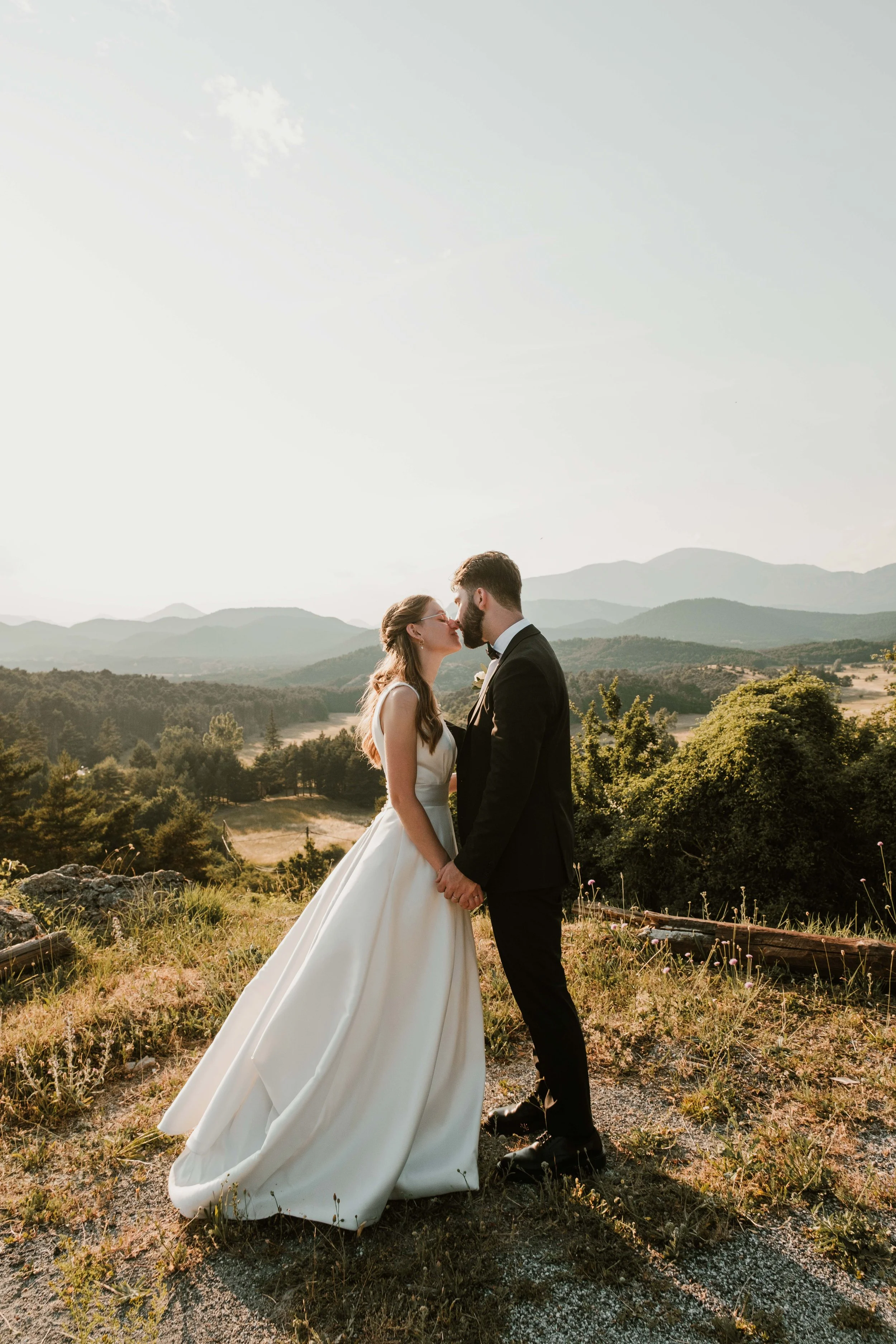 Un couple de mariés en costume et robe de mariée se tient à l'extérieur, s'embrassant avec un paysage de montagnes et de forêts en arrière-plan, sous un ciel clair.