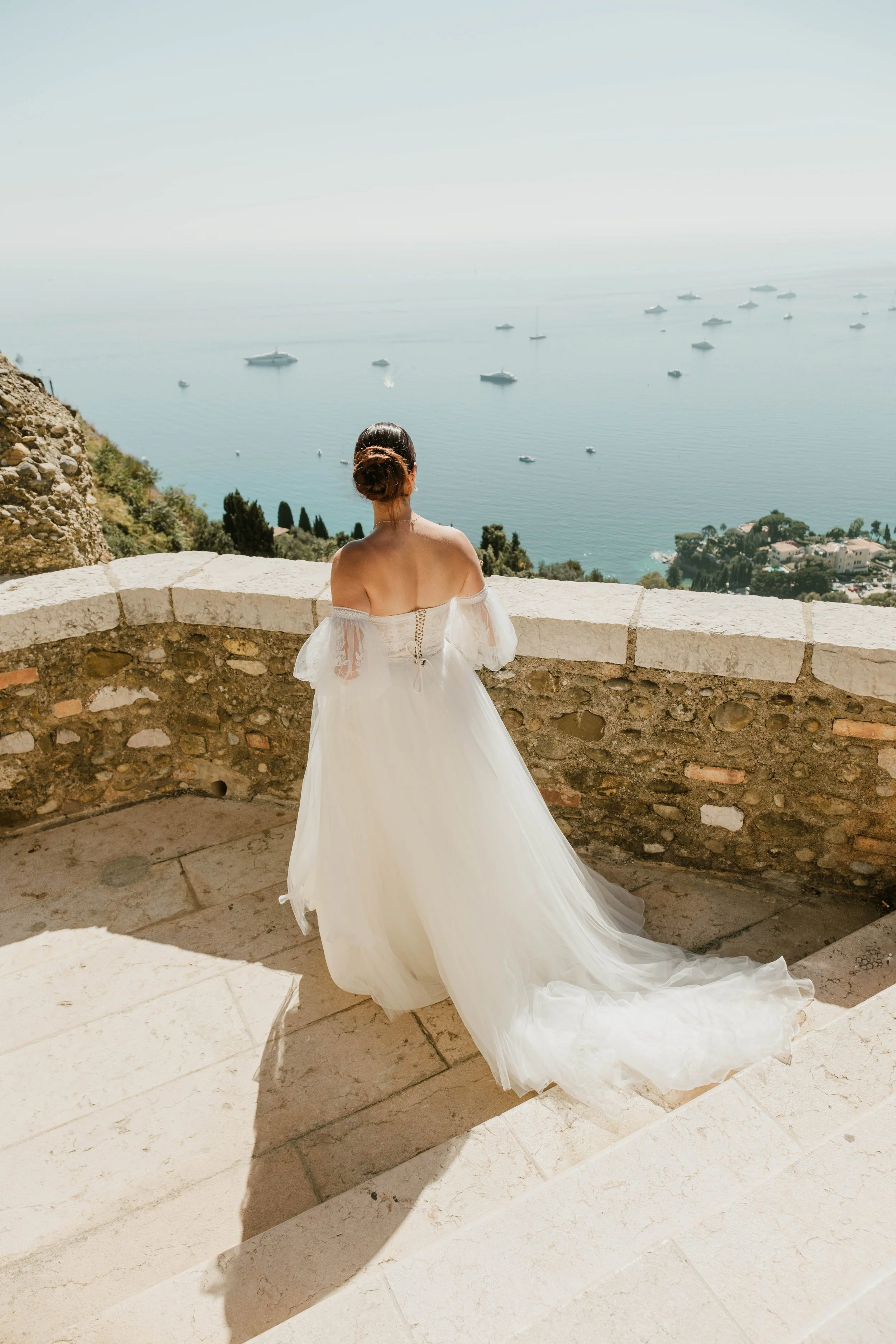 Femme en robe de mariée blanche regardant la mer, sur une terrasse en pierre avec vue sur la côte et plusieurs bateaux à flot.