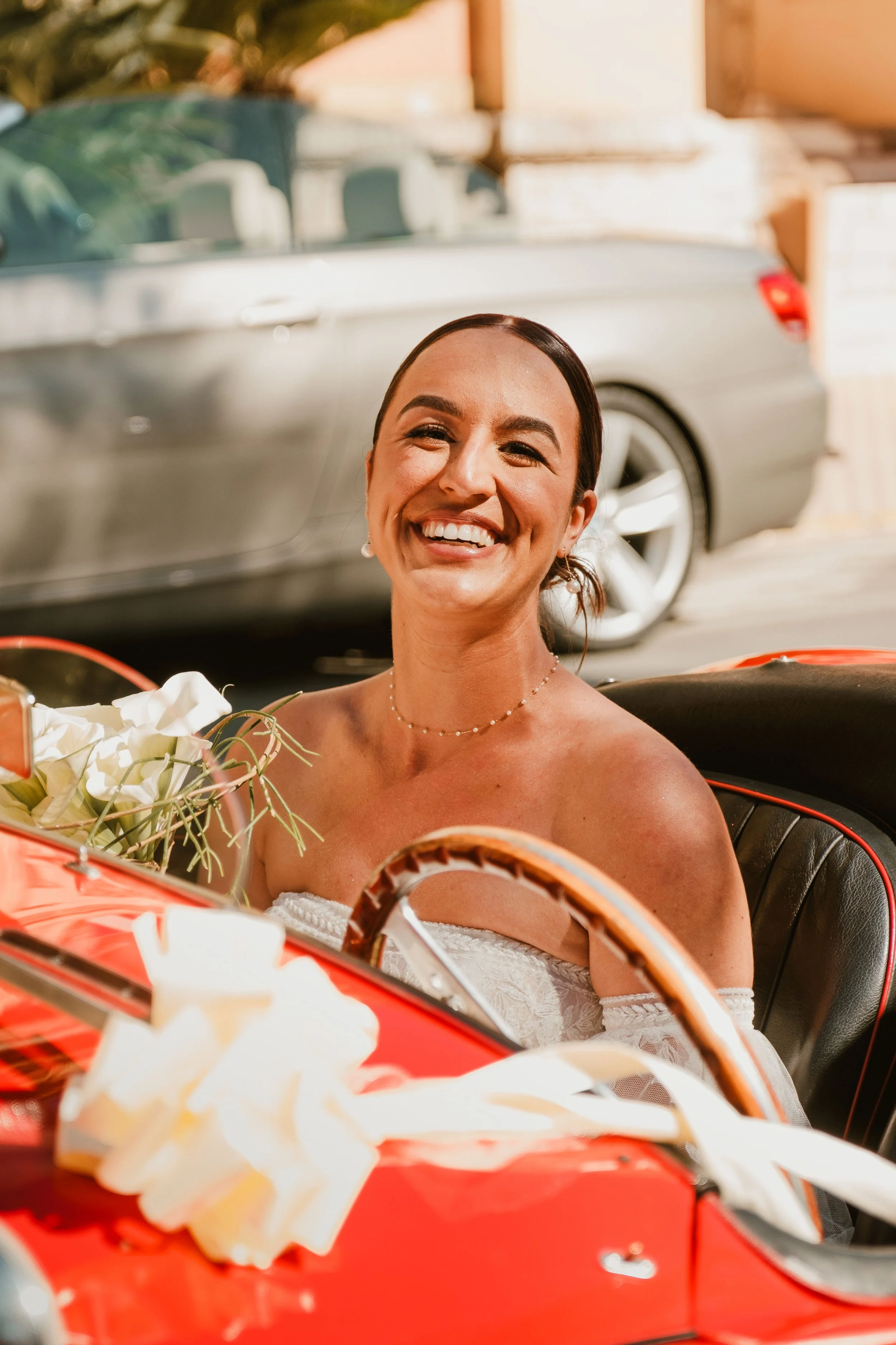 Une femme souriante, en robe blanche, assise dans une petite voiture rouge décorée avec des fleurs