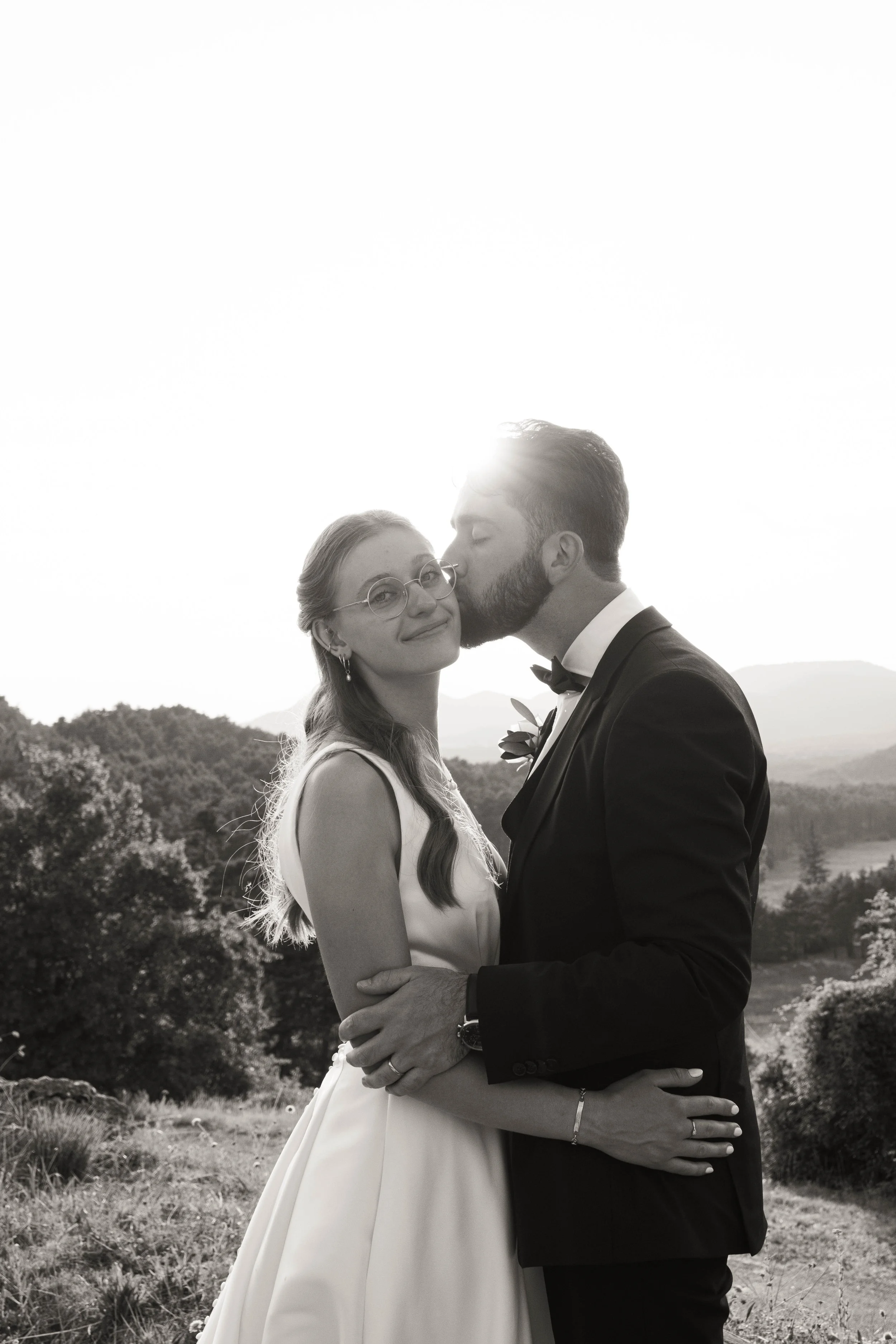 Un couple de mariage pose en extérieur, la femme en robe de mariée et lunettes, l'homme en costume avec nœud papillon, dans un paysage naturel avec des montagnes en arrière-plan.