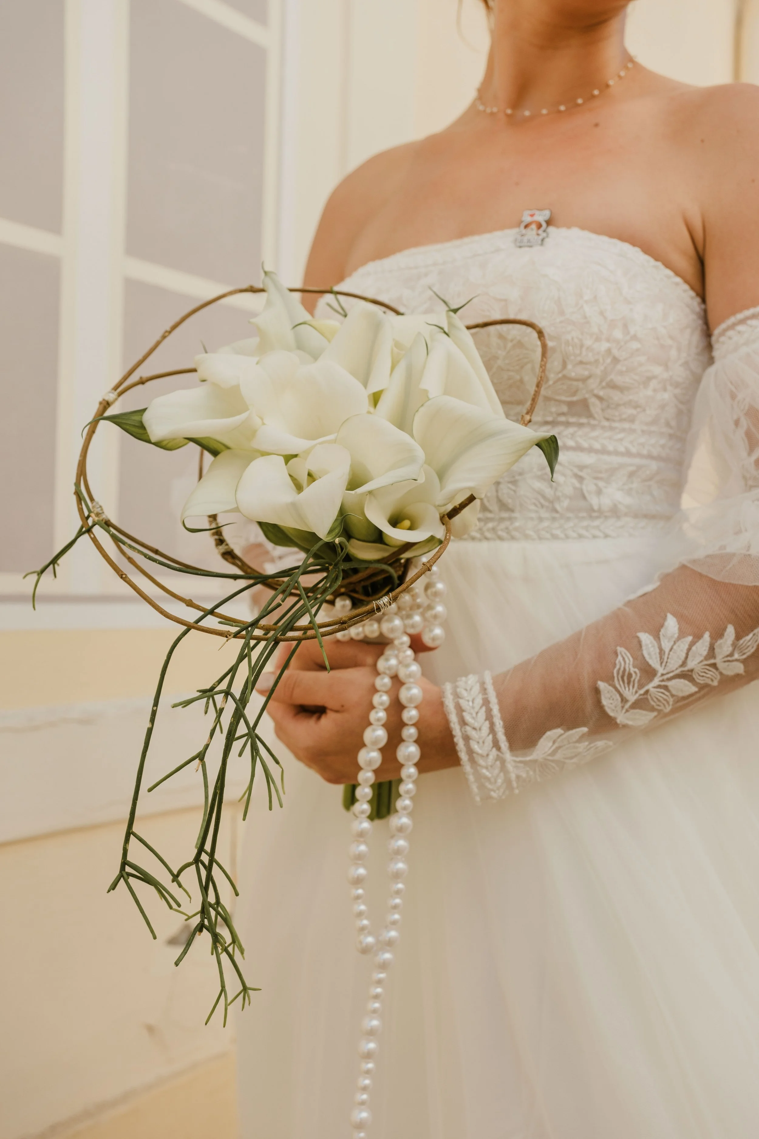Une femme en robe de mariée blanche tient un bouquet de calla lilies, décoré de perles et de tiges de plantes, avec des manches en dentelle et un collier de perles.